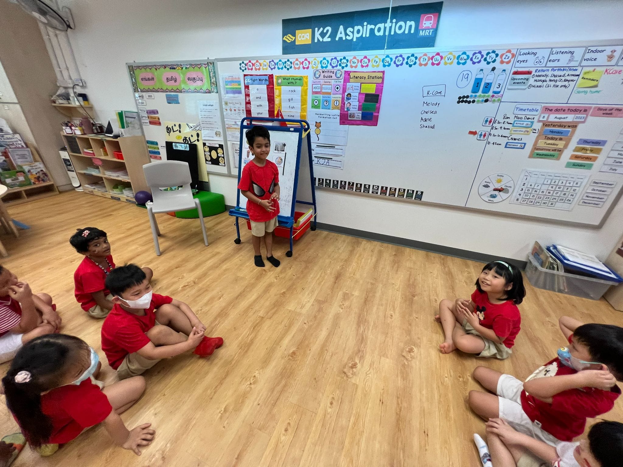 A child in red stands before classmates seated on the floor in a colorful K2 classroom with whiteboards and charts.