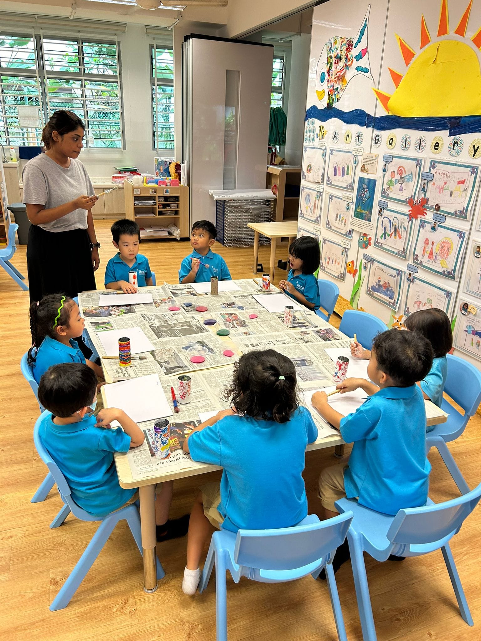 Seven young children in blue uniforms do an art craft activity at a table while a teacher supervises in a colorful classroom.