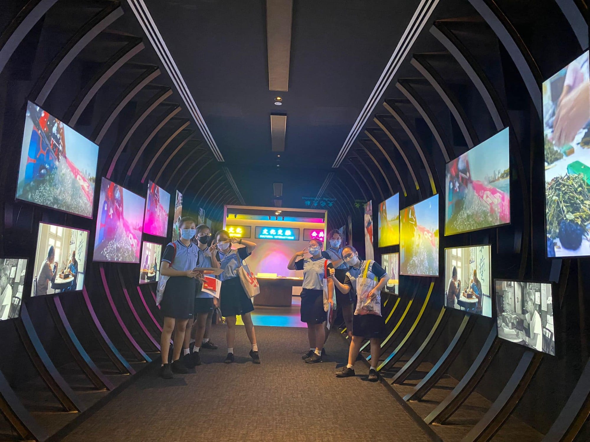 Students in school uniforms and masks pose inside a dark tunnel corridor lined with colorful illuminated display screens.