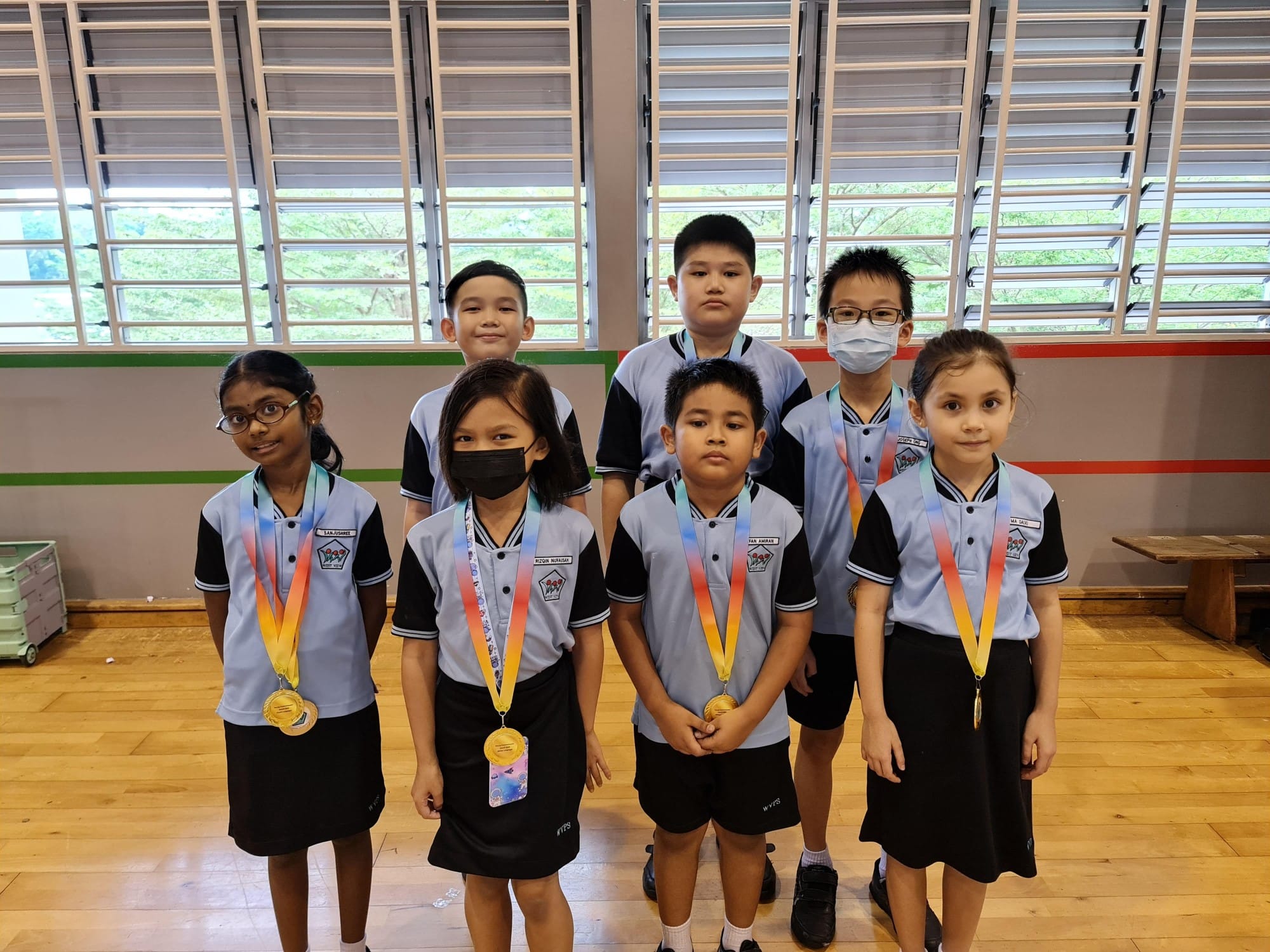 Seven school children in matching uniforms pose indoors, with four wearing gold medals on colorful ribbons.