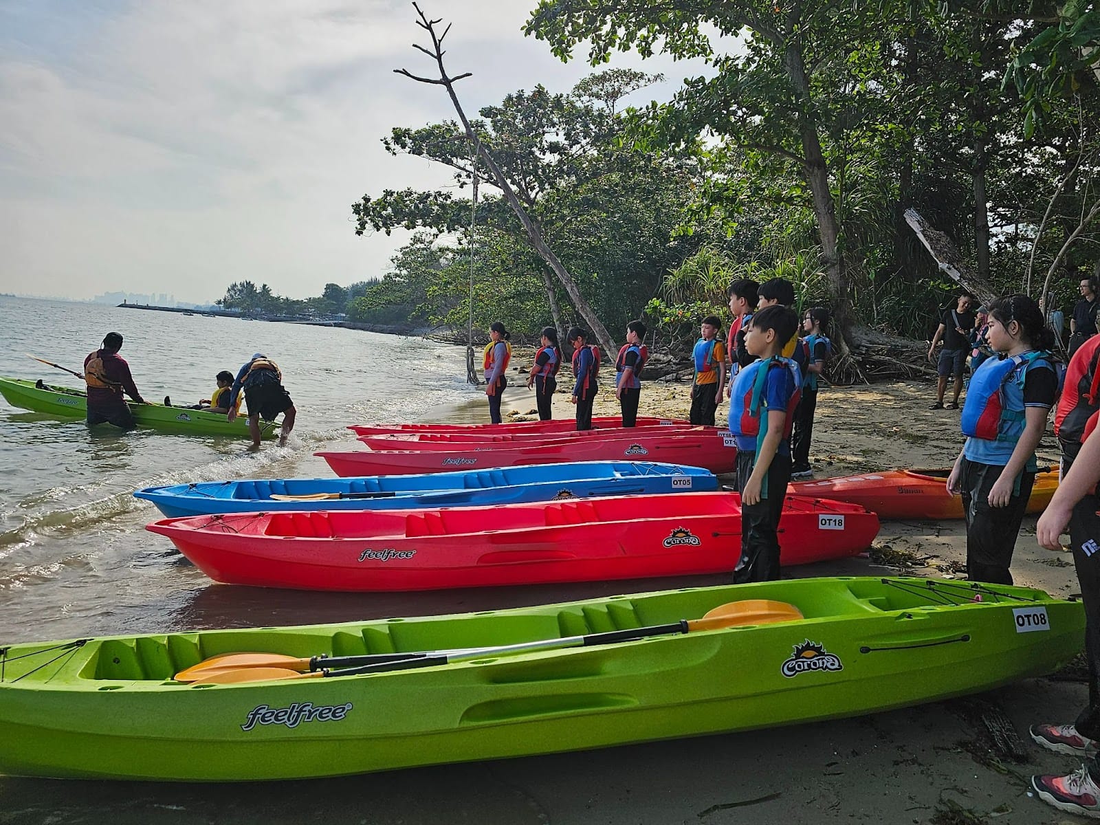 Children in life vests stand on a sandy shore beside rows of red, blue, and green kayaks near calm water.