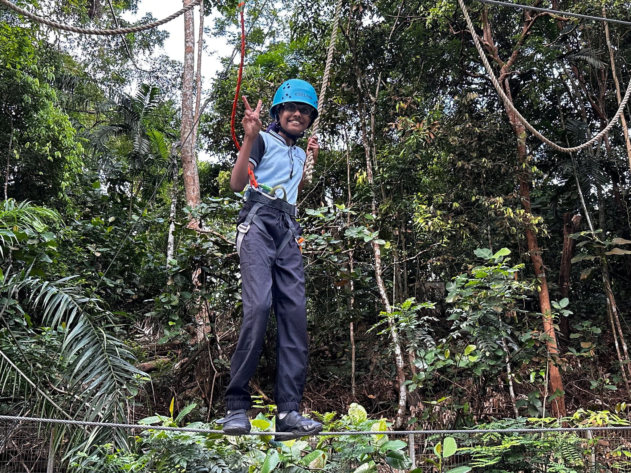 A smiling child in a blue helmet and harness stands on a wire rope course in a lush green forest, flashing a peace sign.