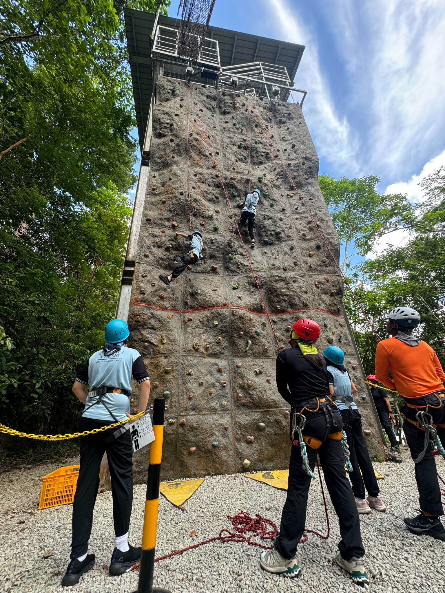 Two people climb a tall outdoor rock wall with harnesses while a group wearing helmets watches from below.