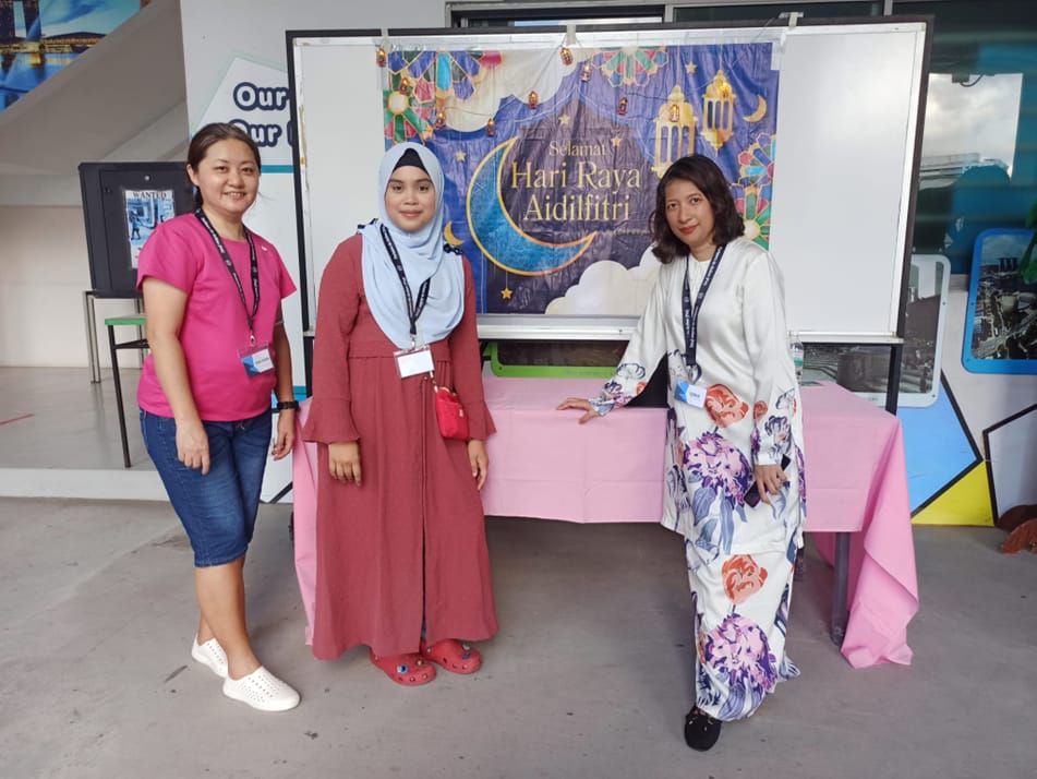 Three women wearing lanyards stand before a Selamat Hari Raya Aidilfitri banner near a pink-clothed table.