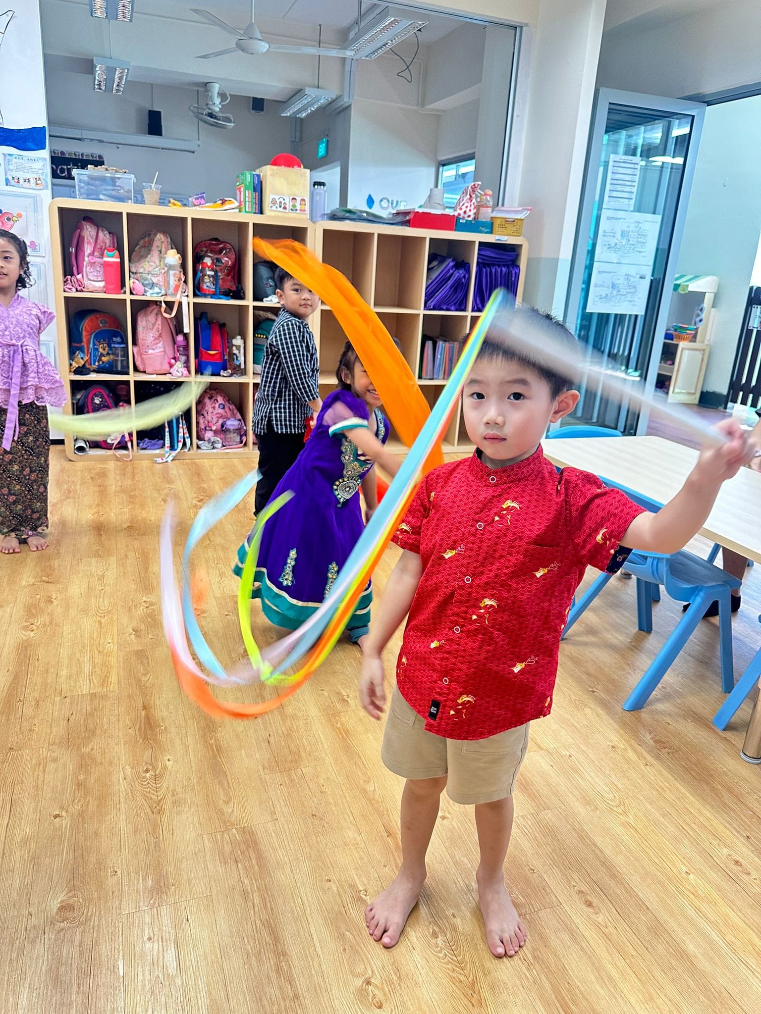 A young boy in a red shirt twirls colorful ribbon wands in a classroom while other children in traditional attire stand nearby.