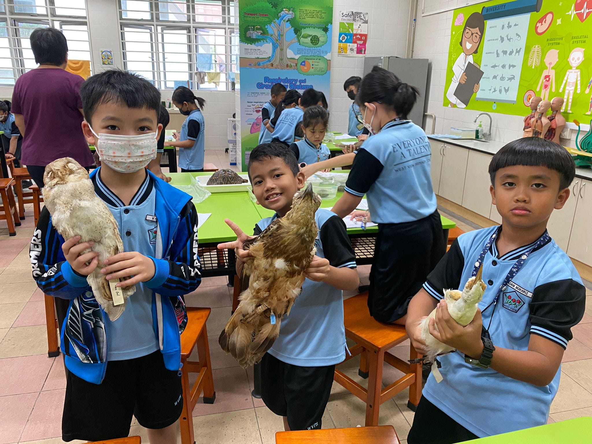 Three young students in blue uniforms hold stuffed birds in a school science classroom with educational posters.