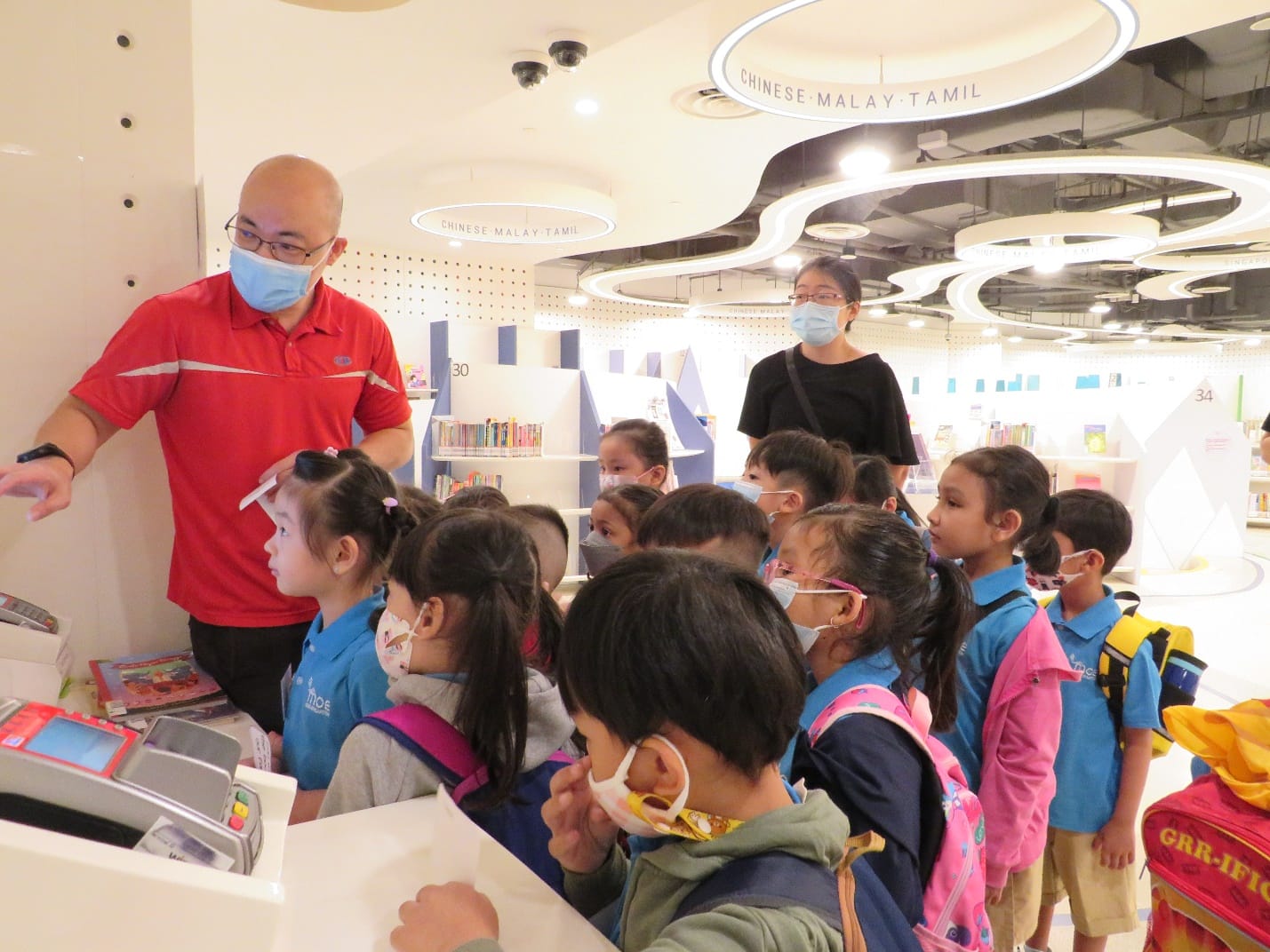 A masked man in red guides a group of masked children in blue uniforms inside a modern library.