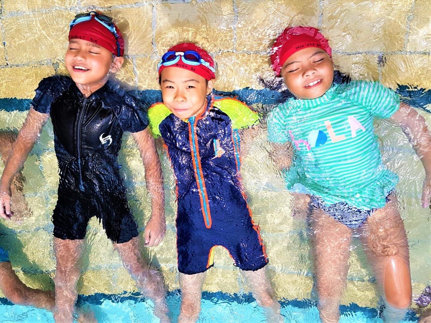 Three smiling children in swimsuits and red swim caps float on their backs in a shallow pool.