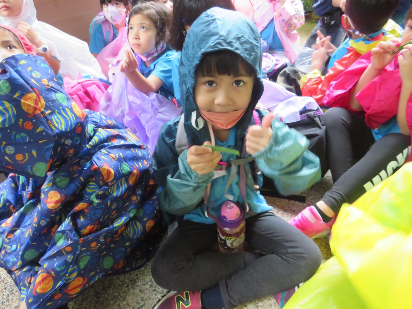 A young girl in a blue hooded jacket and pink face mask sits cross-legged among other children with colorful rain gear.