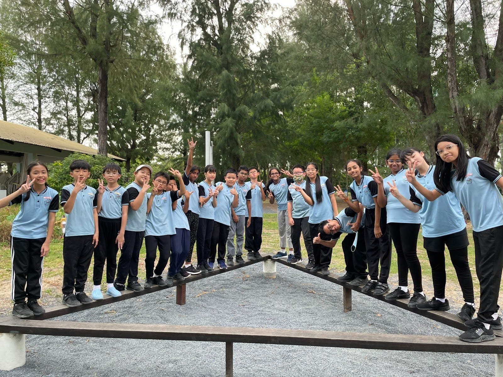 A group of students in light blue uniforms stand on a V-shaped wooden balance beam outdoors, posing with peace signs.