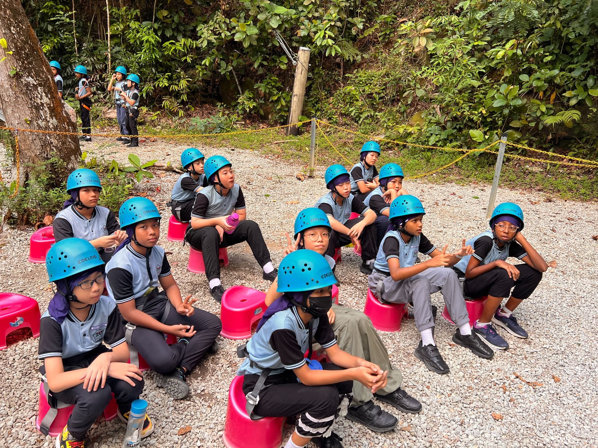 Students in blue helmets and school uniforms sit on pink stools on gravel near a forest during an outdoor activity.
