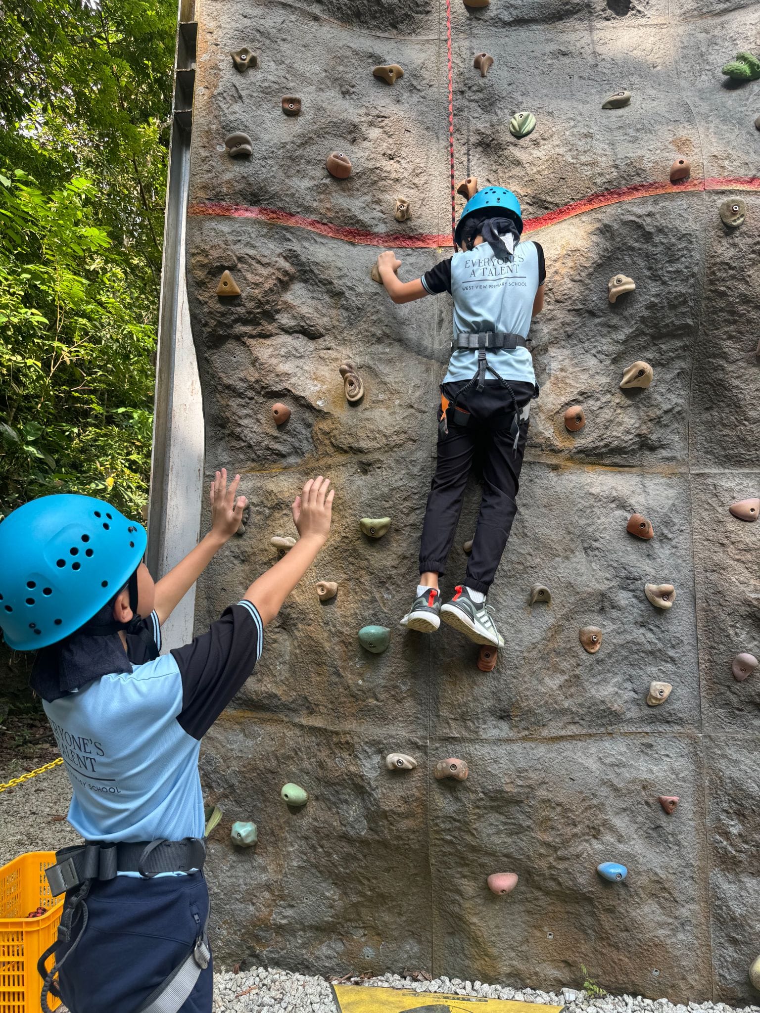 Two children in blue helmets and harnesses at an outdoor climbing wall, one climbing up while the other watches.