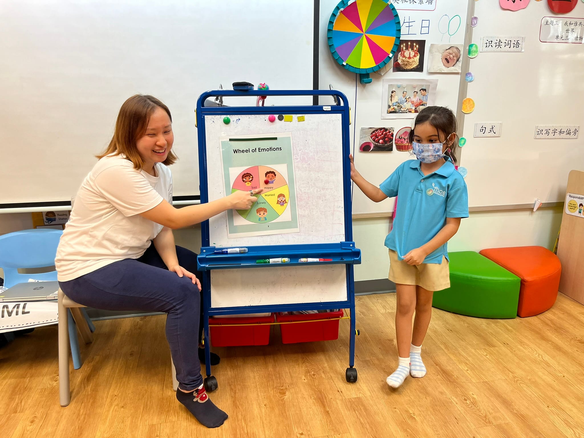 A smiling teacher points to a Wheel of Emotions chart on an easel beside a masked young girl in a classroom.