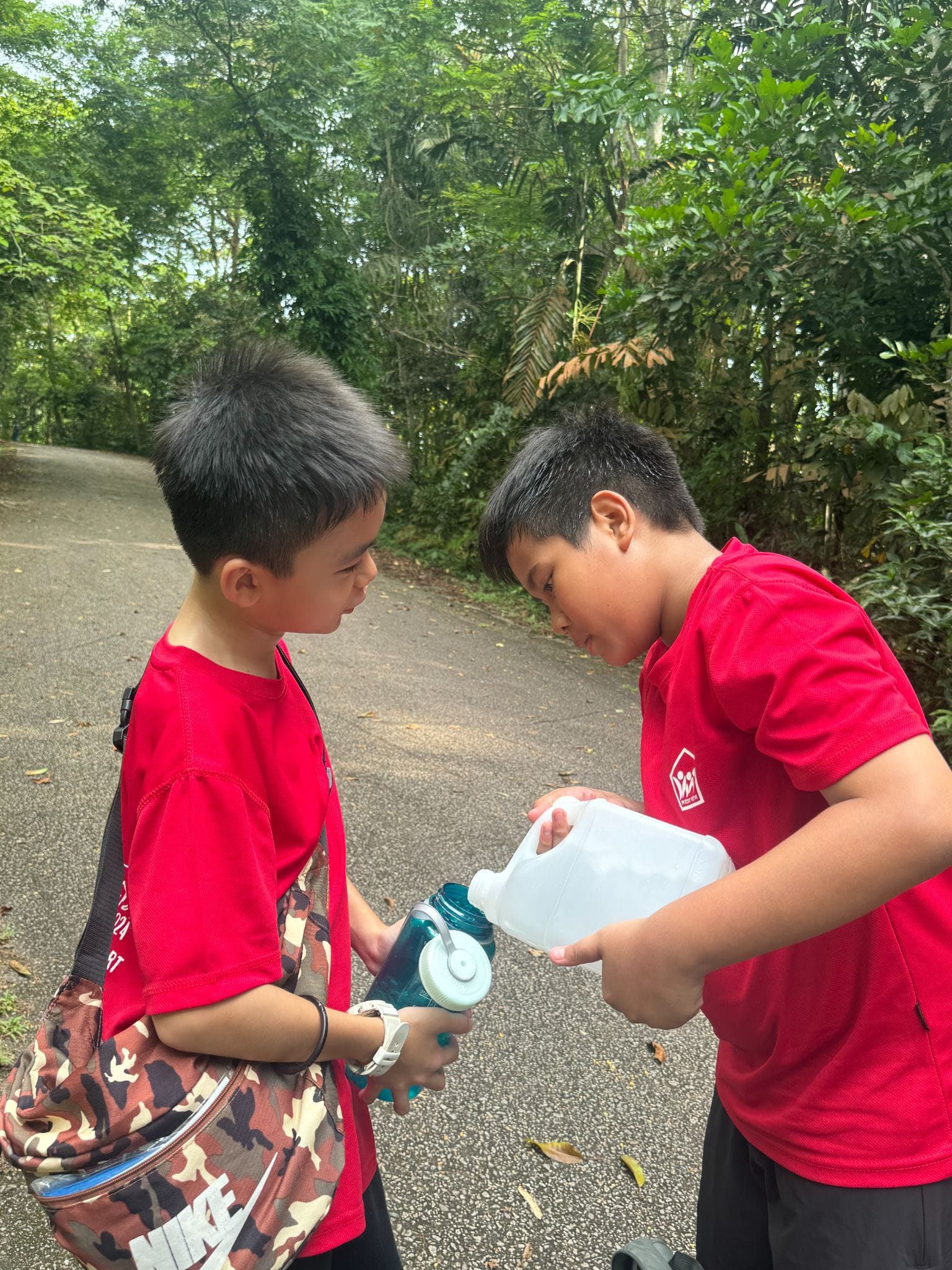 Two boys in red shirts on a tree-lined path, one pouring water from a jug into the other's bottle.