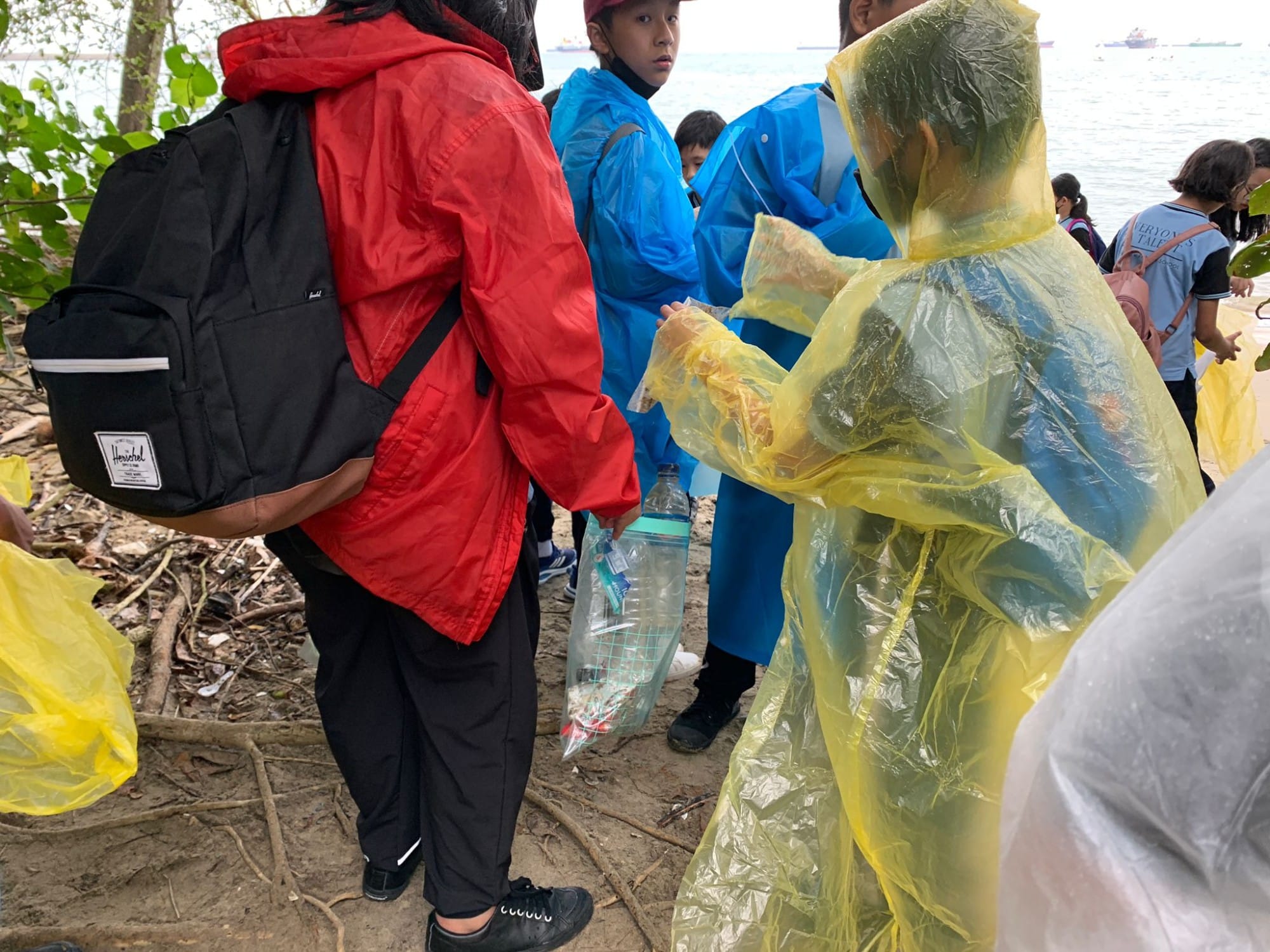 Children in rain ponchos collect plastic bottles and trash on a muddy shoreline near water.