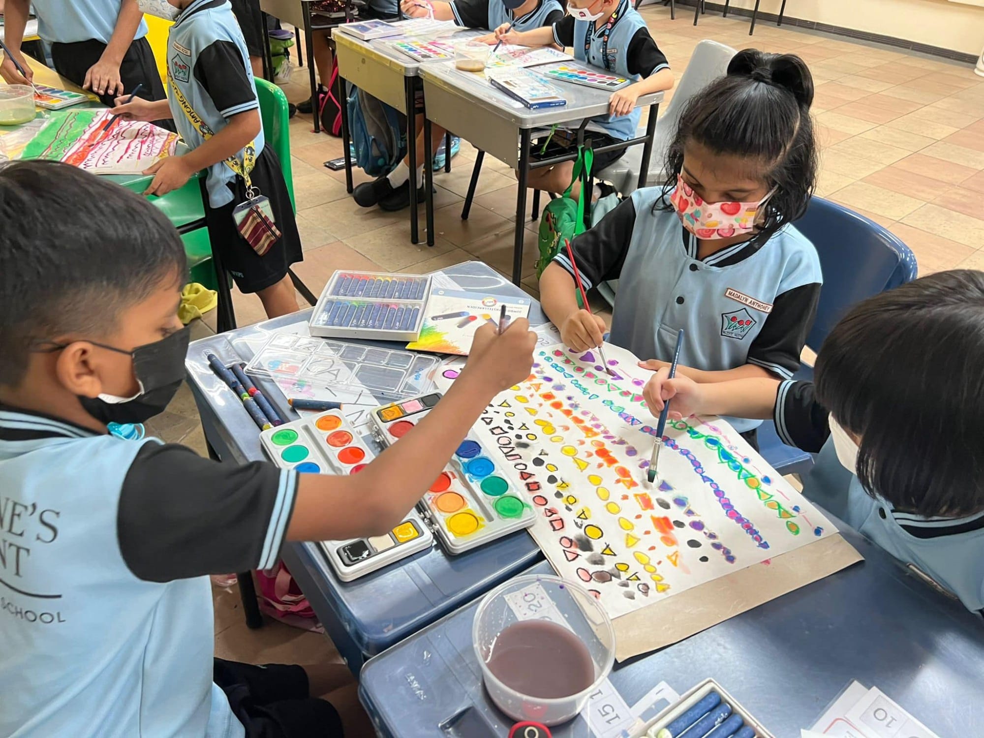 Three masked schoolchildren in blue uniforms paint colorful shapes on a large sheet using watercolors at a classroom desk.