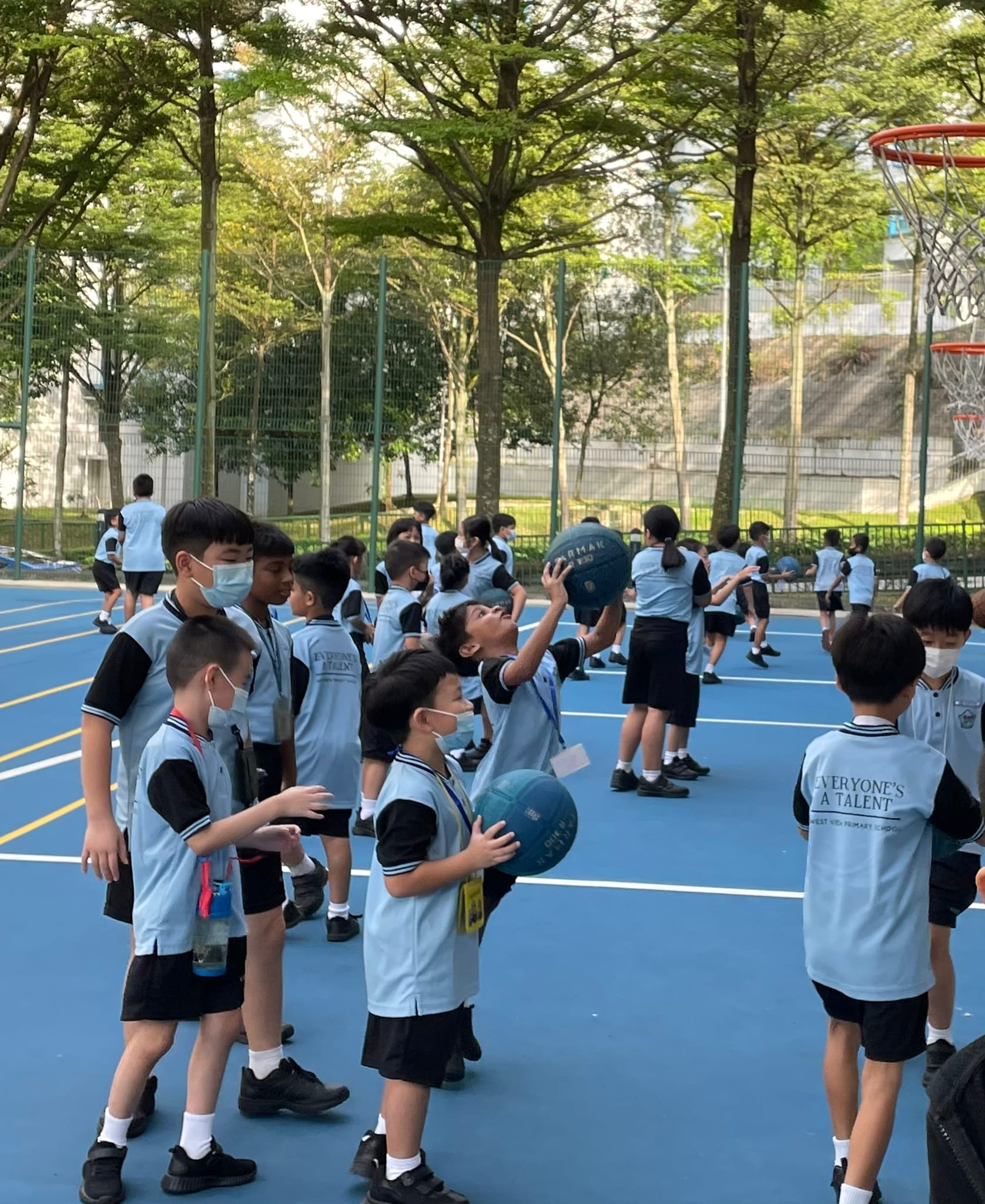 Children in school uniforms and face masks play with basketballs on an outdoor blue court surrounded by trees.