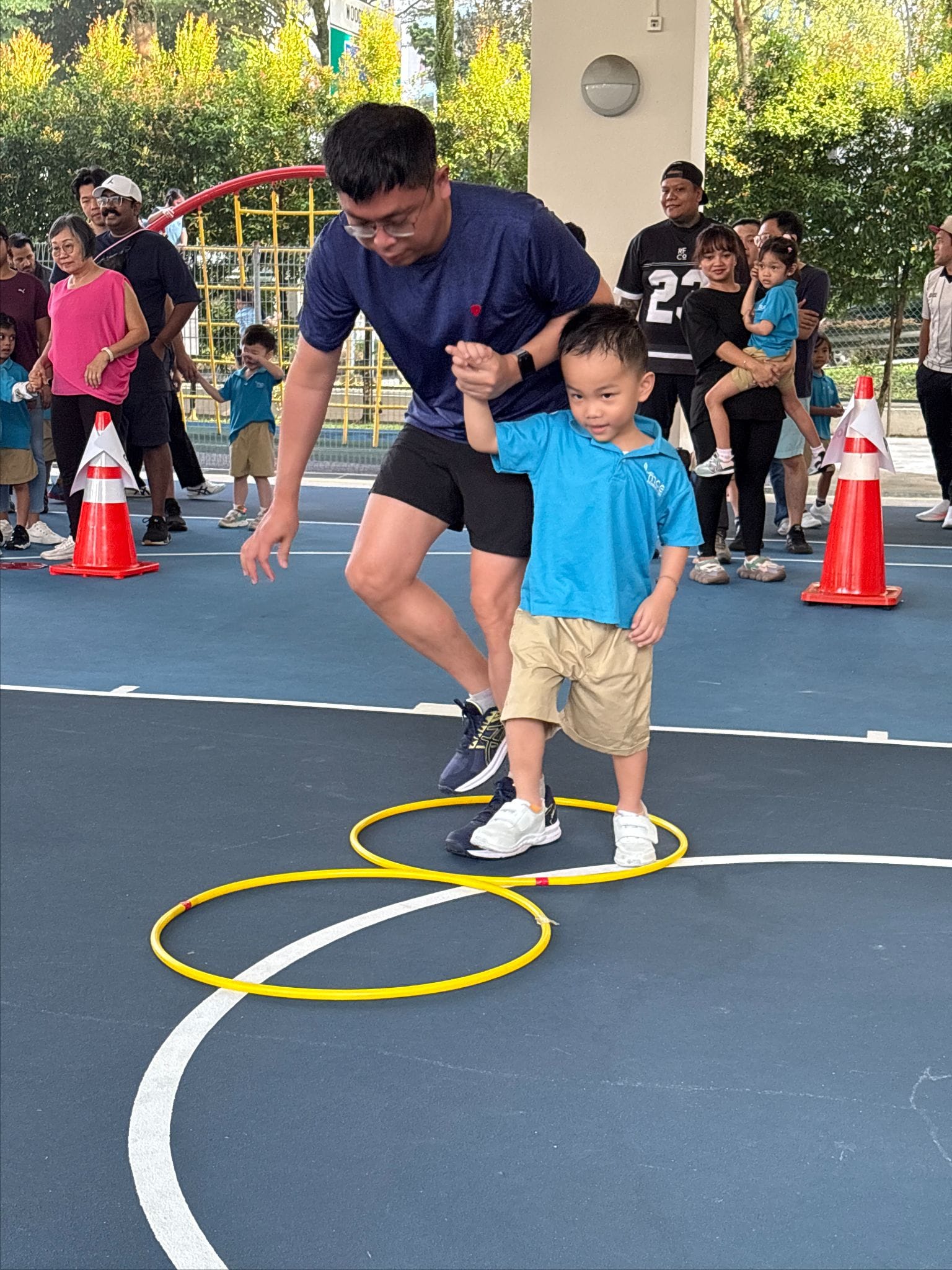 A man guides a young boy stepping through yellow hula hoops on an outdoor court, with spectators watching nearby.