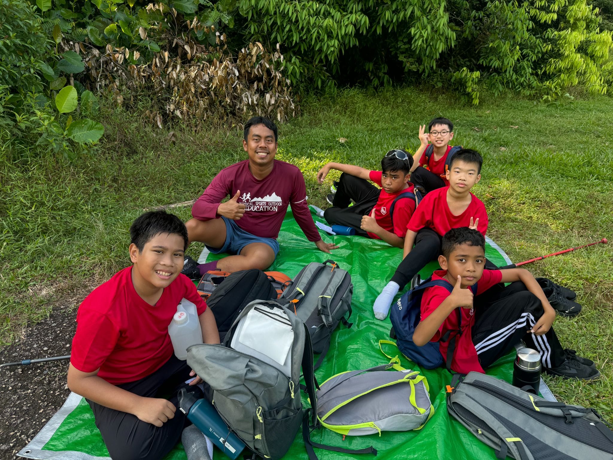Six boys in red shirts and an adult sit on a green tarp outdoors with backpacks, smiling at the camera.