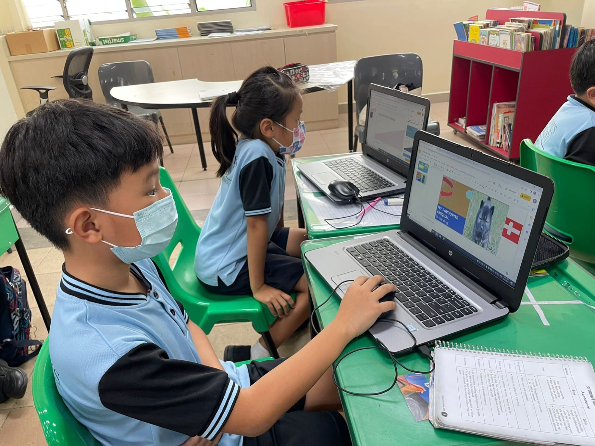 Two masked children in school uniforms sit at green desks using laptops in a classroom with a bookshelf nearby.