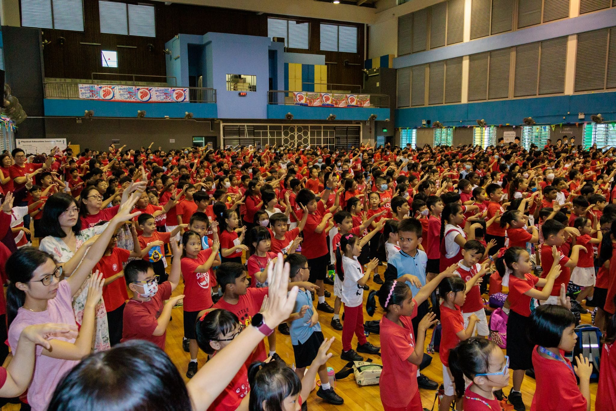 Hundreds of children in red shirts raise their hands during a group activity in a large school hall.