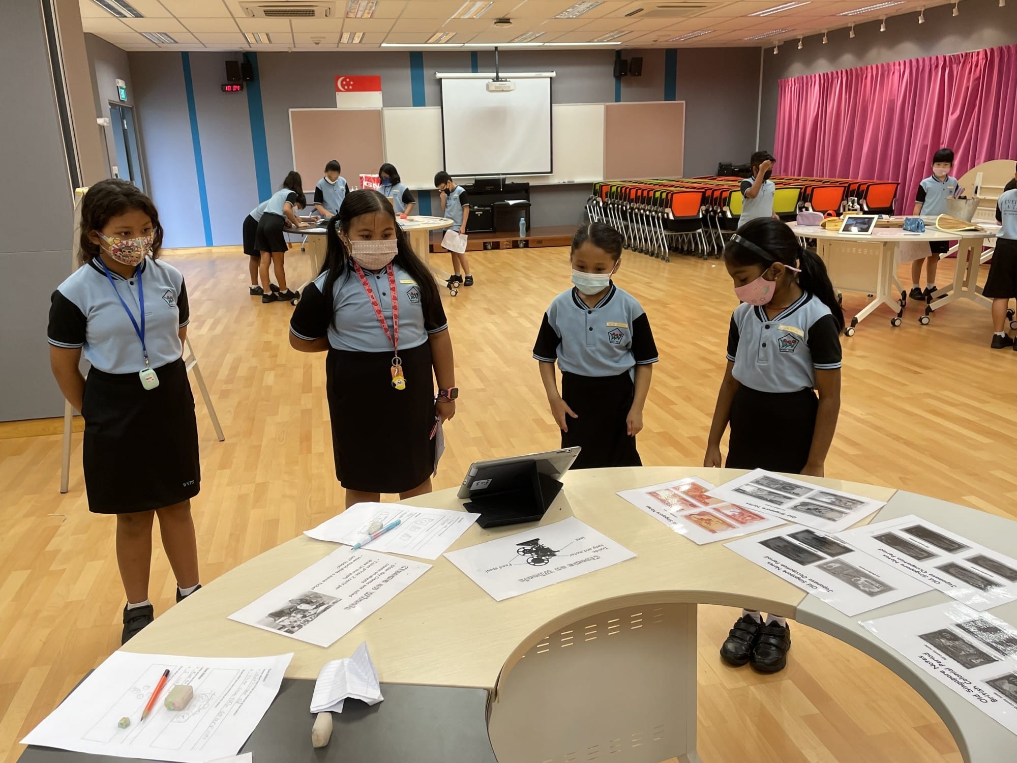 Four masked schoolgirls in blue uniforms stand around a table with papers and a tablet in a school hall.