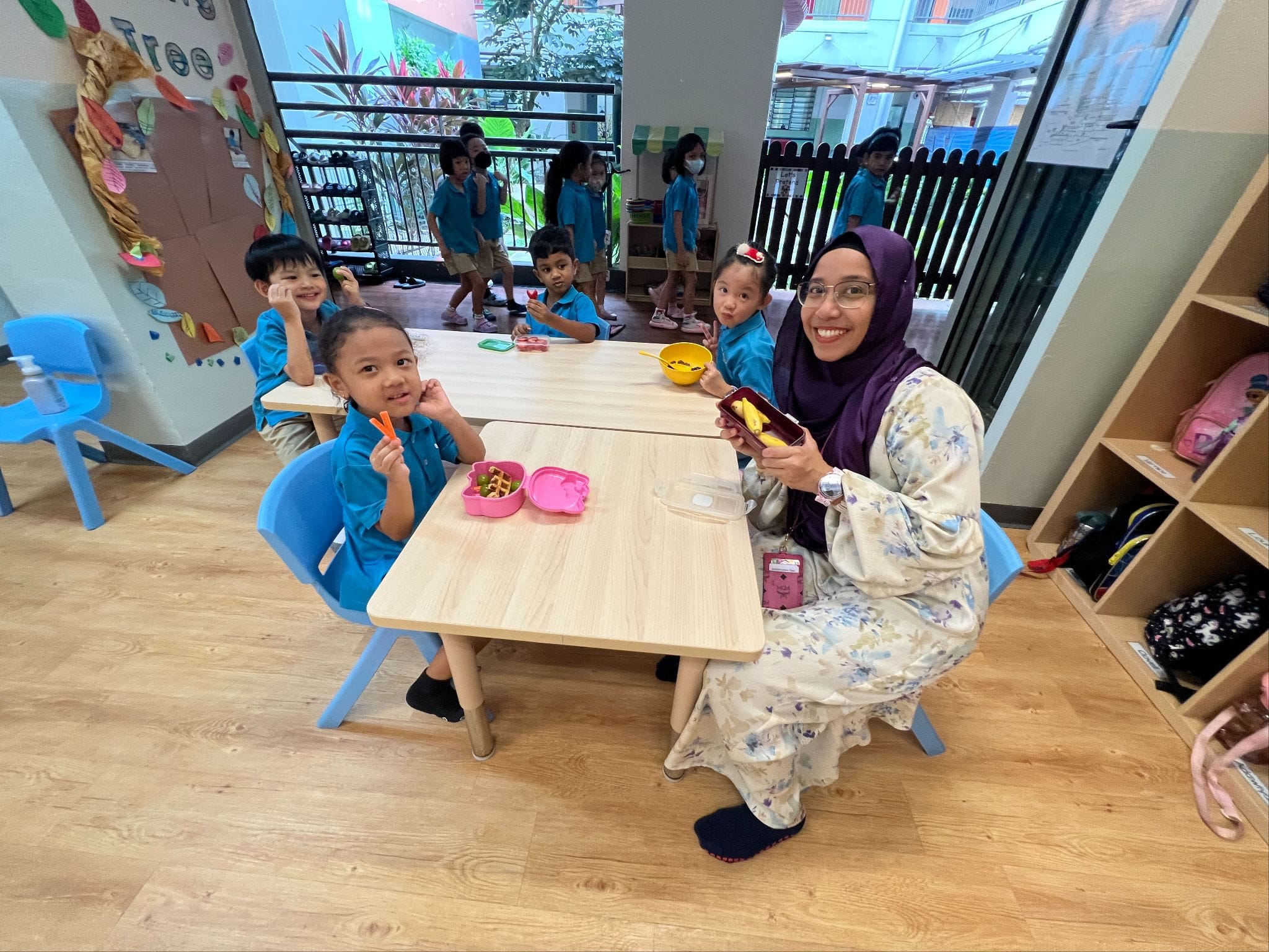 A smiling teacher in a hijab sits at a low table with young children in blue uniforms eating snacks in a bright classroom.