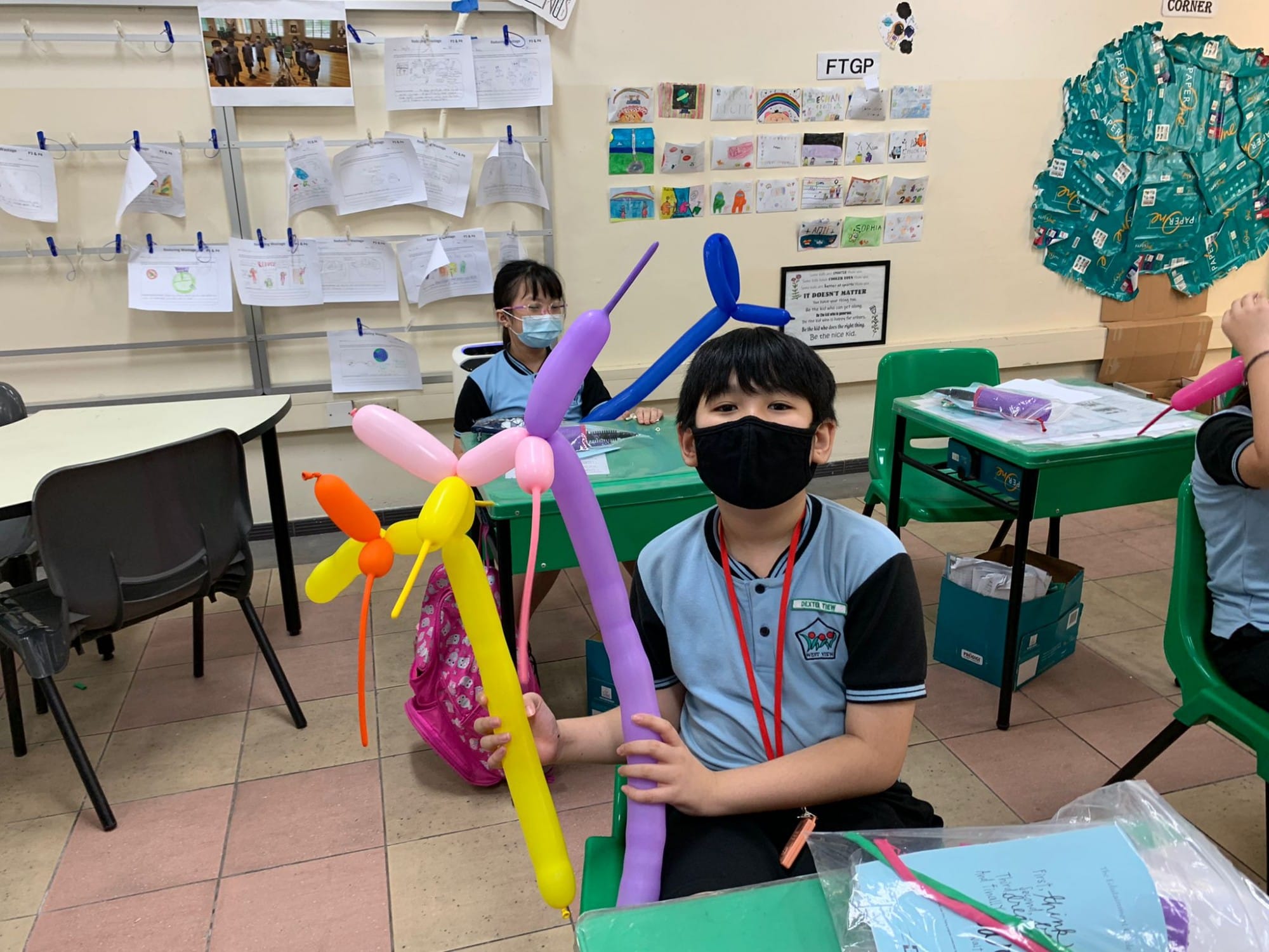 A masked child in a classroom holds colorful twisted balloon sculptures in purple, yellow, pink, and blue.