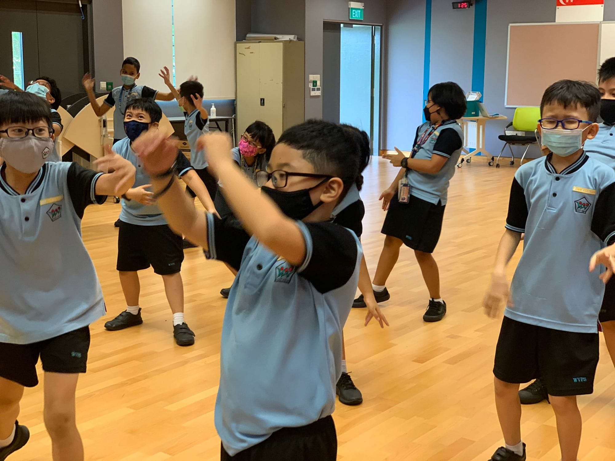 Children in light blue school uniforms and face masks dancing or exercising in an indoor hall with wooden floors.