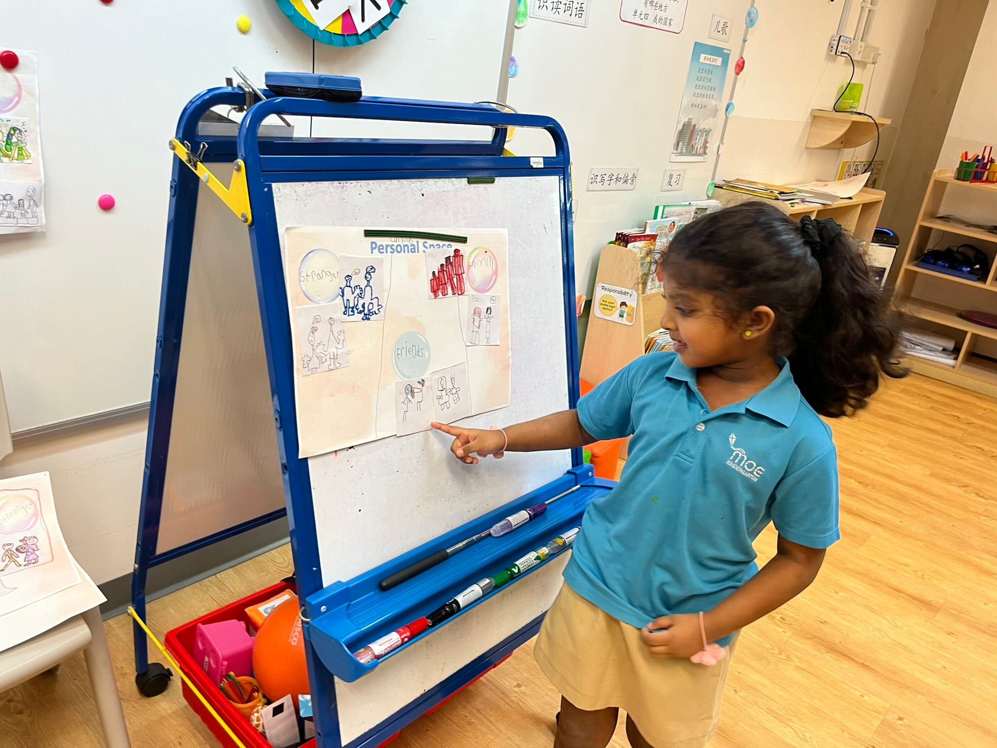Young girl in blue uniform pointing at a "Personal Space" poster on a classroom easel.