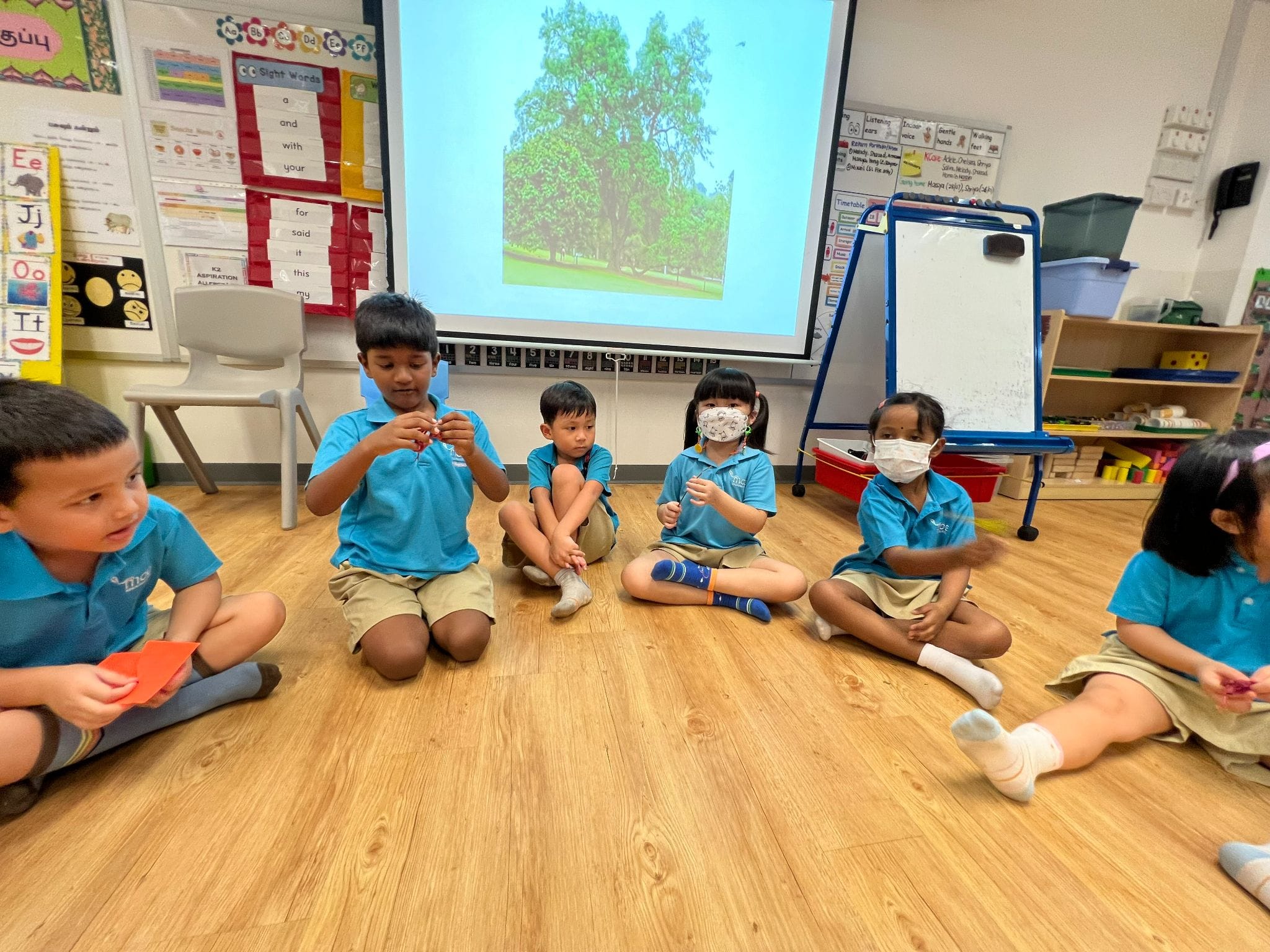 Five young children in blue uniforms sit on a classroom floor, with a projected tree slide and alphabet charts behind them.