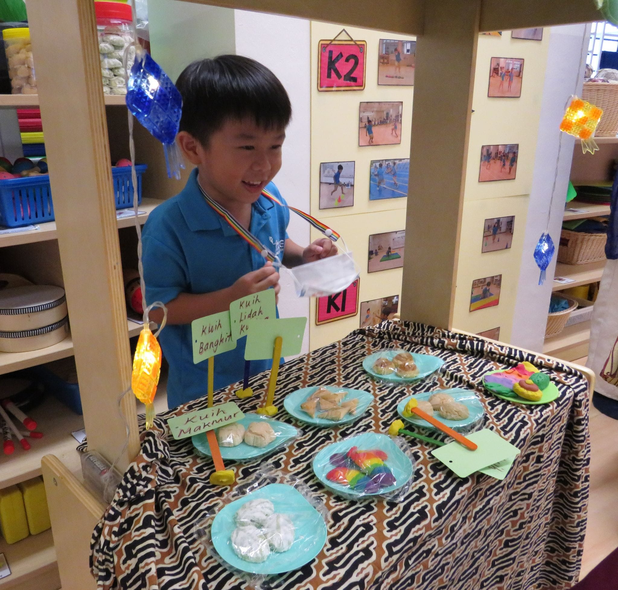 A smiling boy in a blue shirt stands behind a play market stall with labeled traditional kuih on plates.