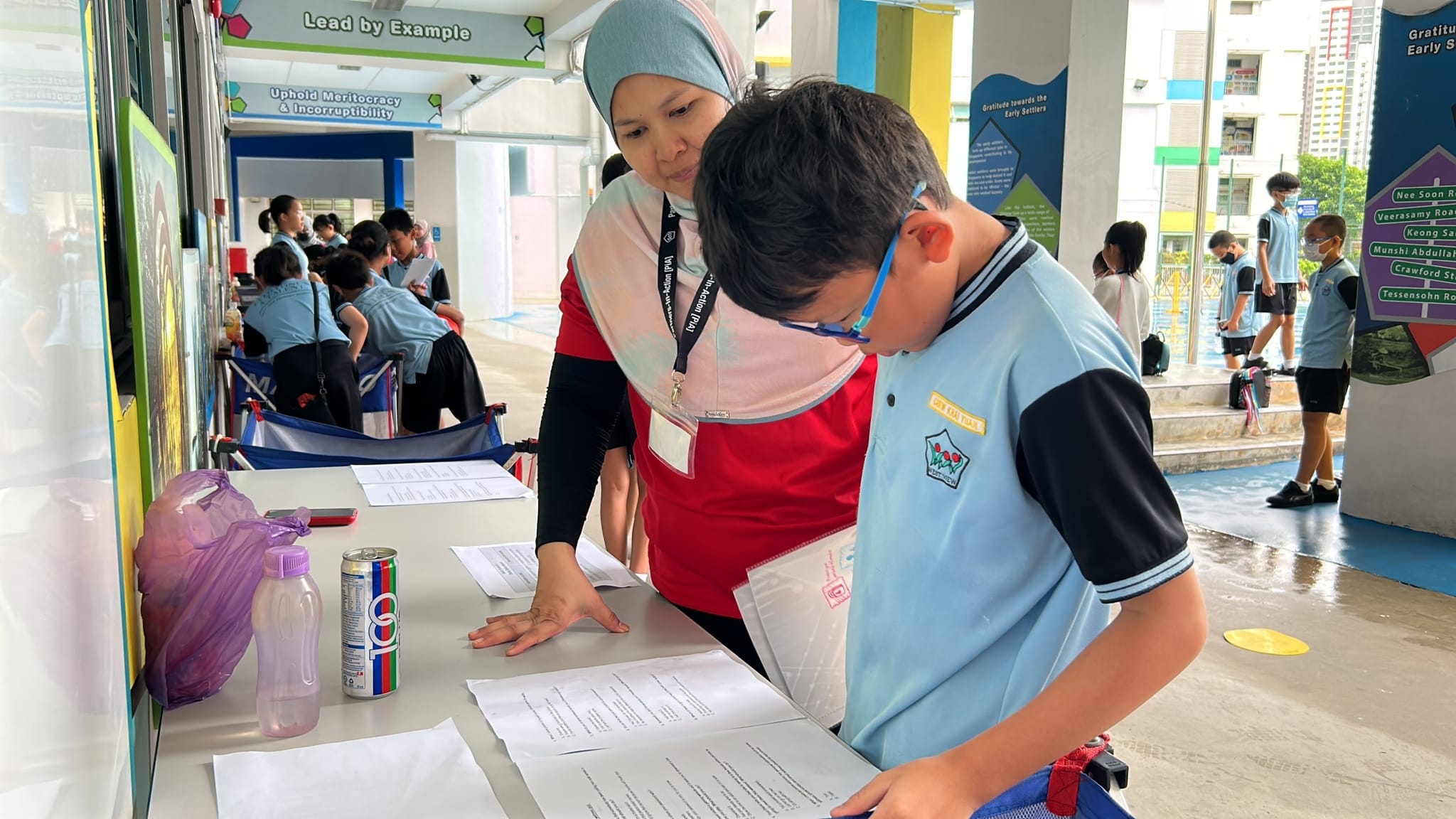 A woman in a red top and hijab leans over to help a young boy in a blue uniform review papers at a table.