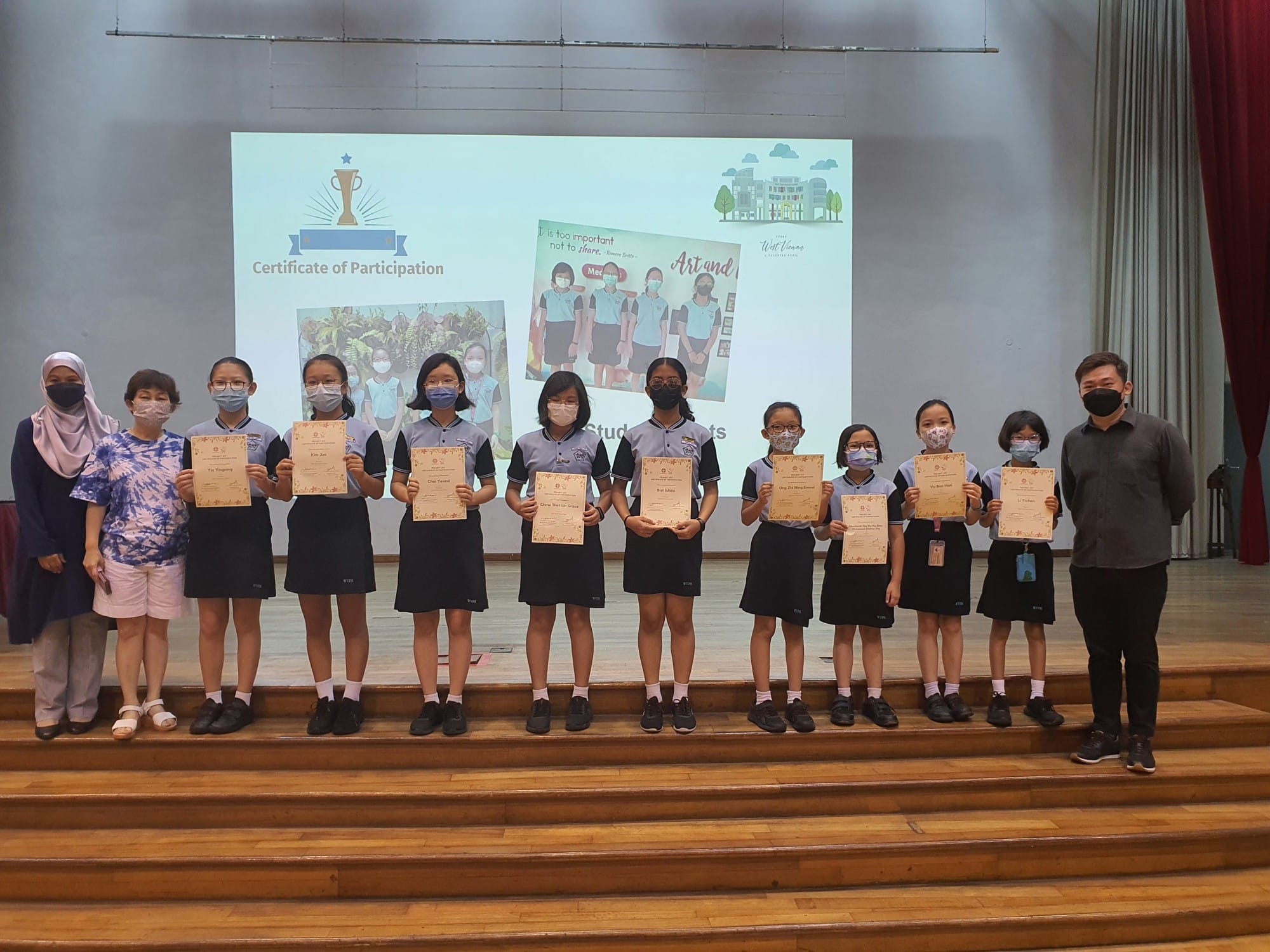 Students in school uniforms and masks hold Certificates of Participation on a stage with a projected screen.
