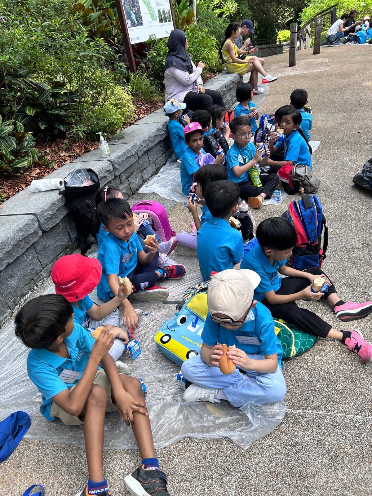 A group of young children in blue uniforms sitting on a path outdoors, eating snacks during a field trip.