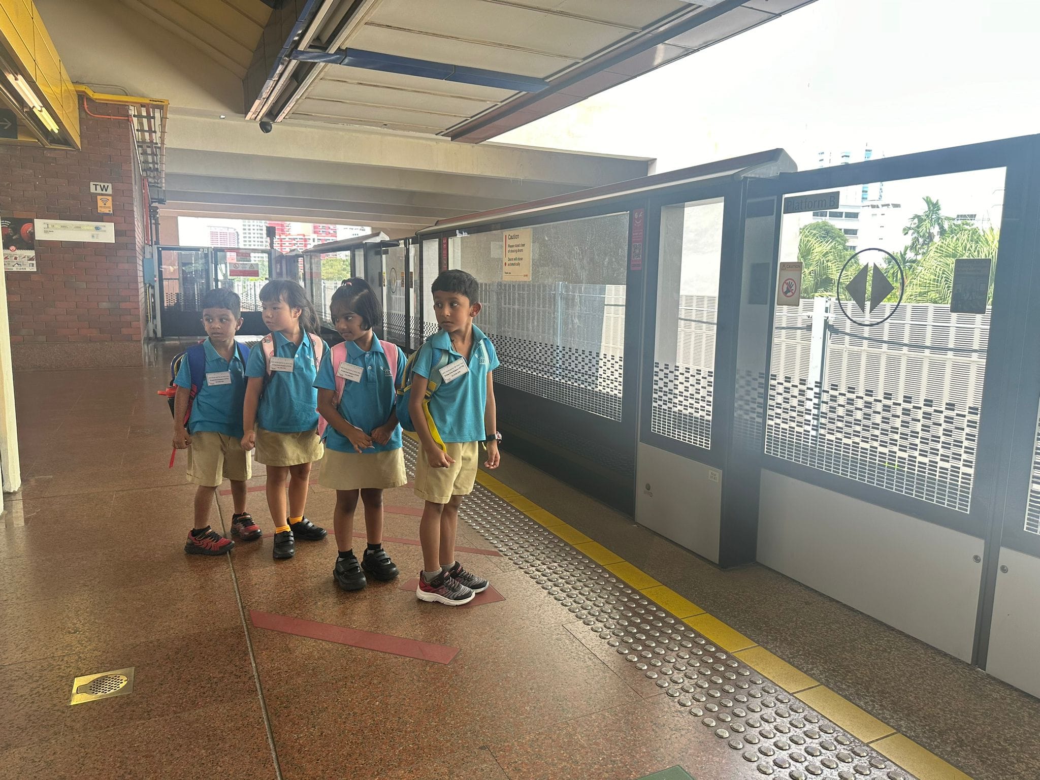 Four young children in blue uniforms and backpacks stand waiting at a train station platform door.