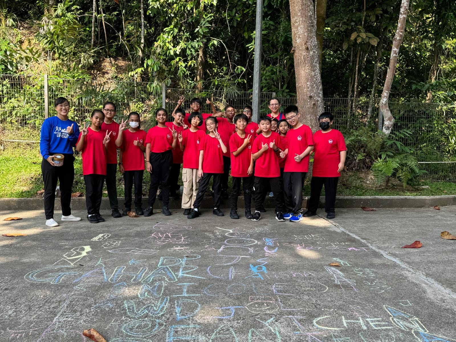 Group of children in red shirts posing outdoors near colorful chalk drawings on pavement, with trees behind them.