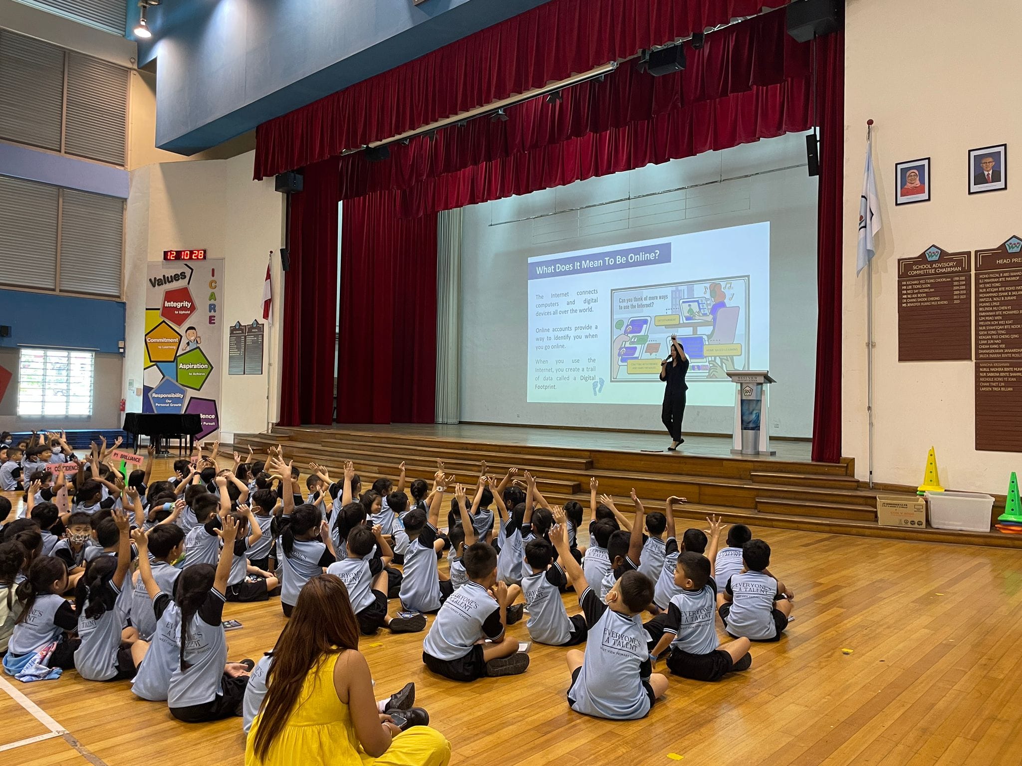 Children in matching shirts sit on a gym floor with hands raised, facing a presenter on stage with an internet safety slideshow.