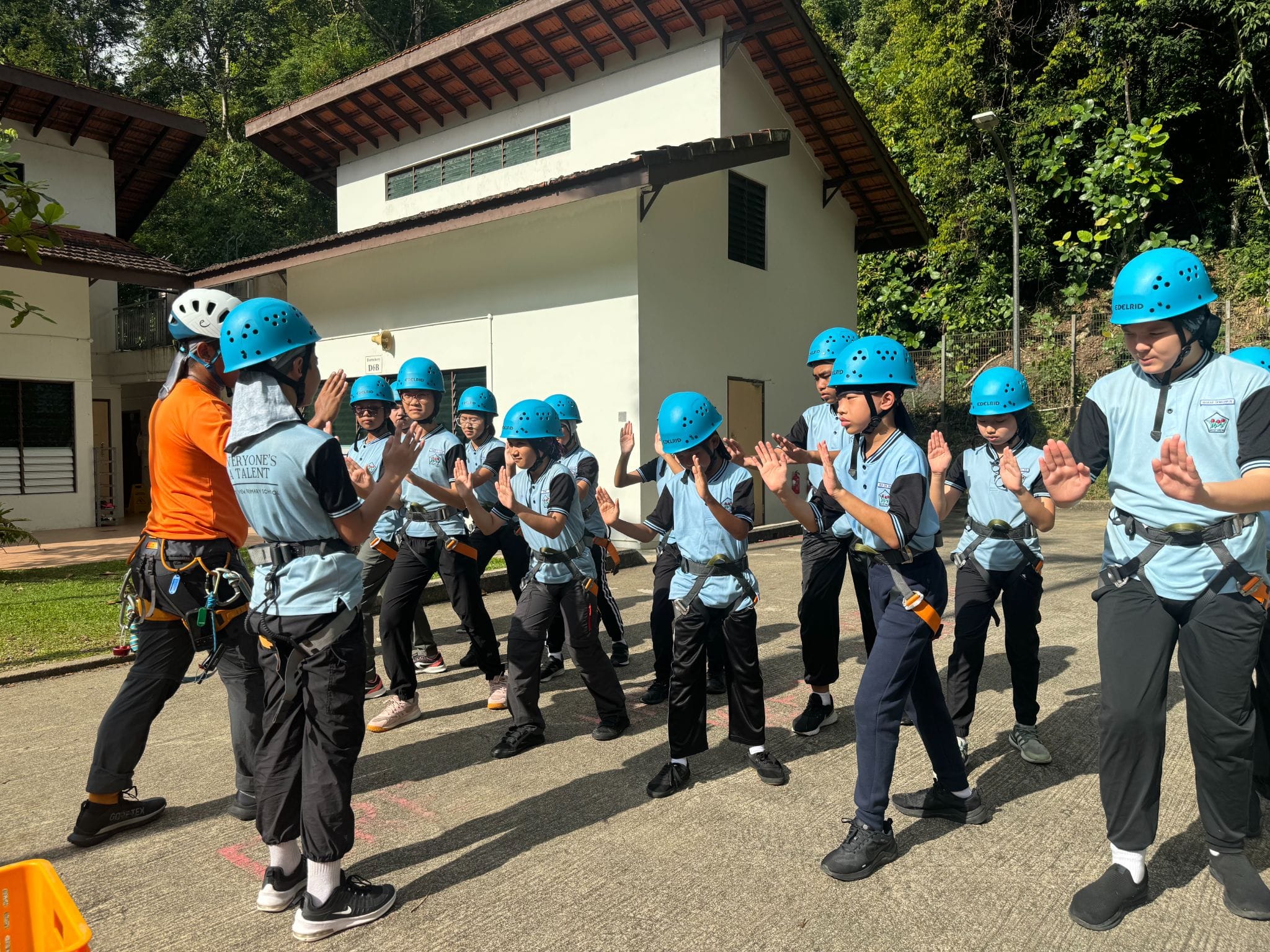 A group of students in blue helmets and harnesses raise their hands during an outdoor activity briefing near a building.