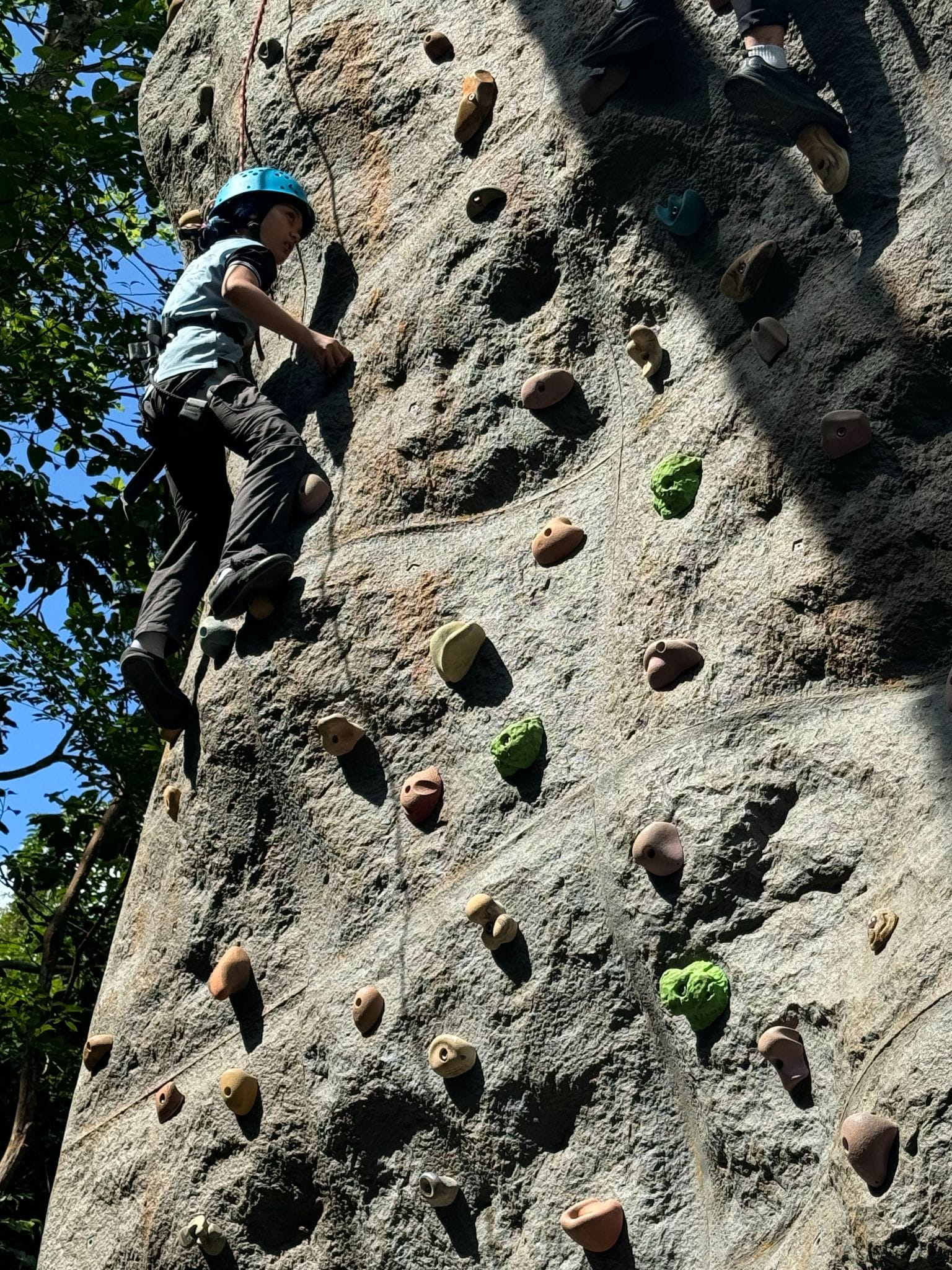 Child wearing a blue helmet and harness climbing a tall rock wall with colorful holds outdoors.