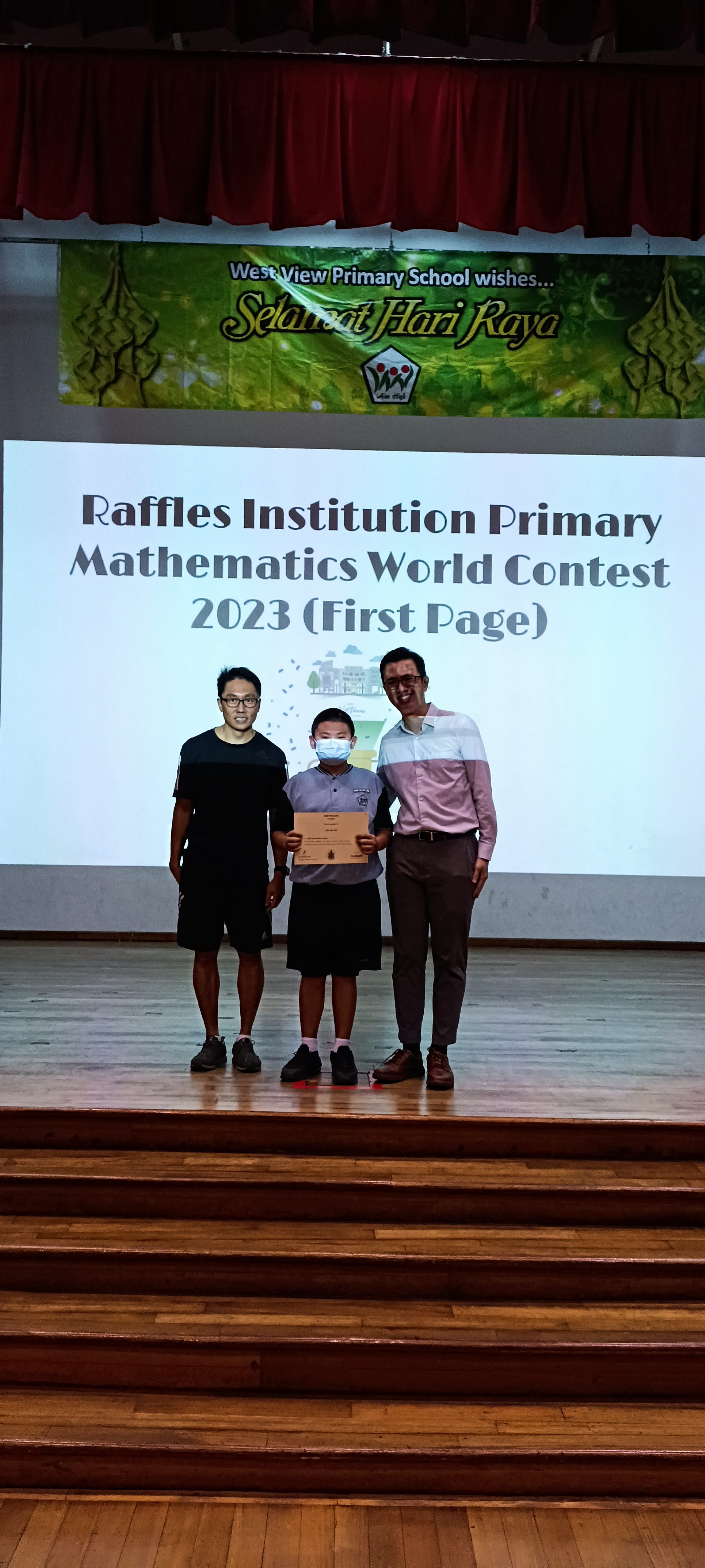 A masked student holds a certificate on stage flanked by two adults at a 2023 Raffles Institution Primary Mathematics World Contest.