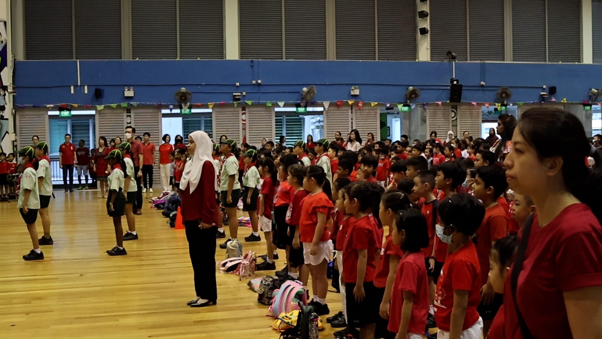 Large group of schoolchildren in red uniforms standing in rows inside a decorated indoor hall with teachers present.