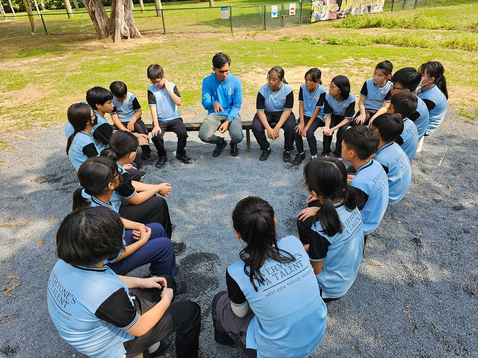 A teacher in blue speaks to students in light blue uniforms seated in a circle outdoors on a gravel area.