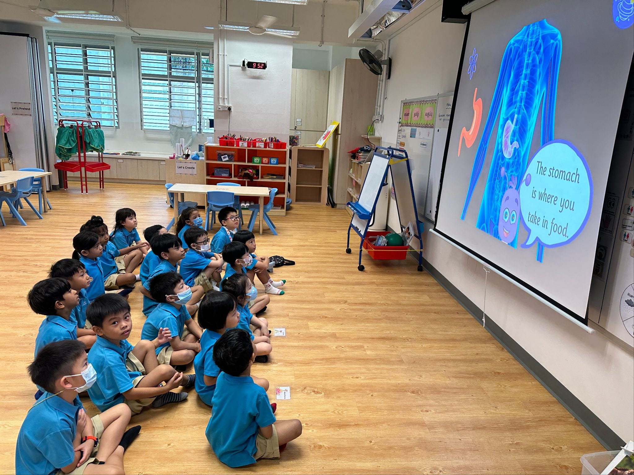 Young children in blue uniforms sit on a classroom floor watching a projected slide about the stomach.