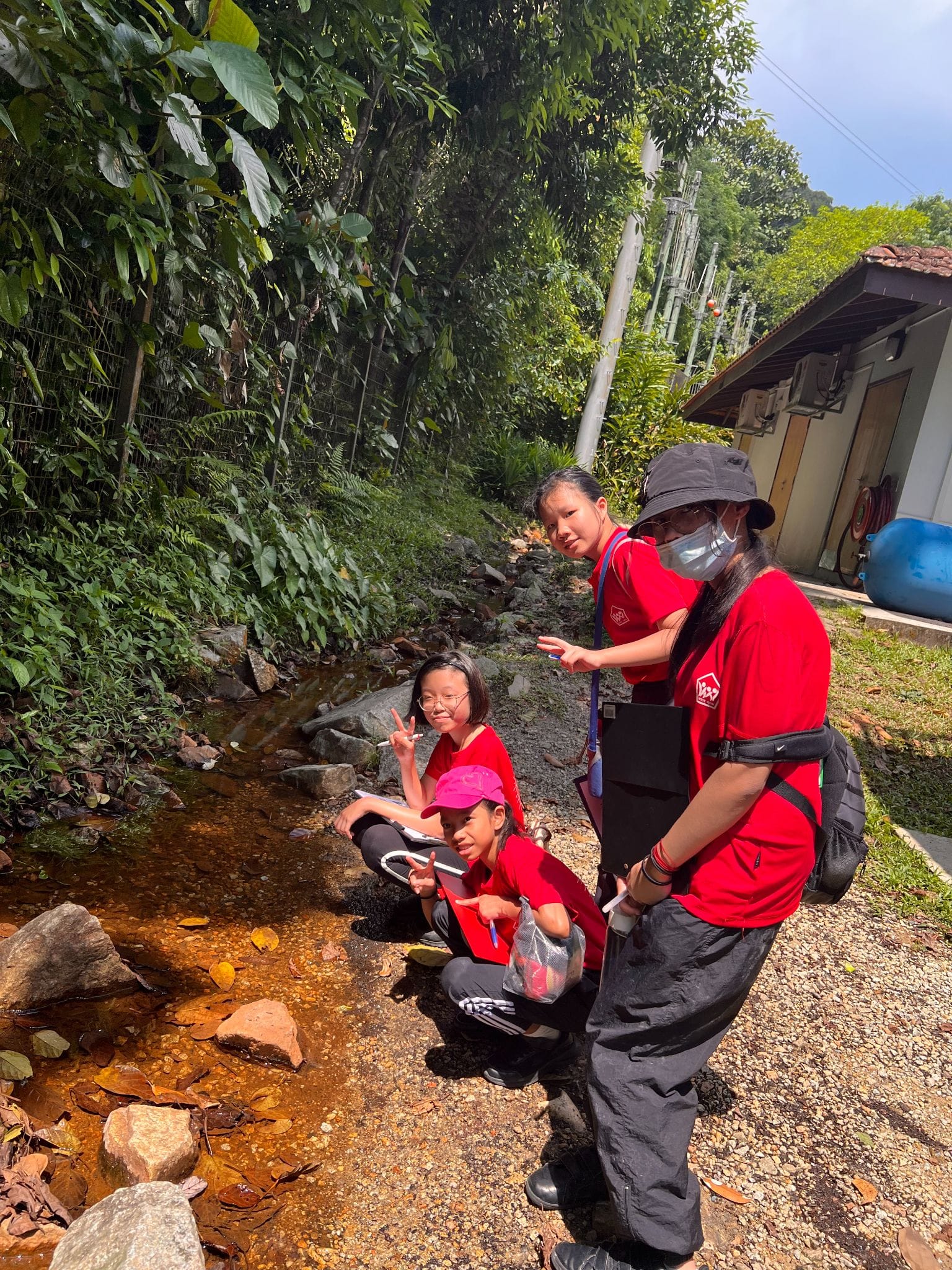 Three people in red shirts crouch beside a rocky stream surrounded by green trees outdoors.