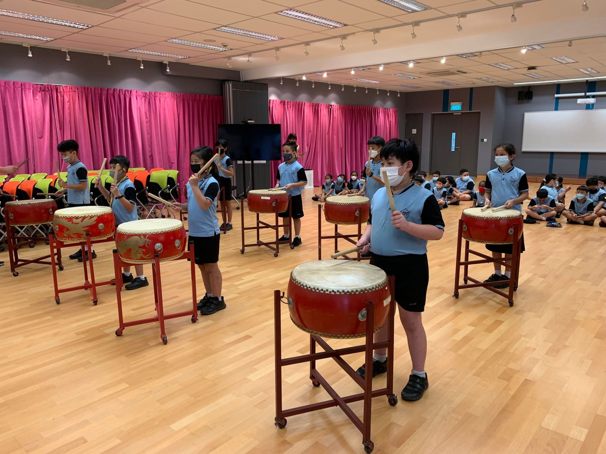 Children in school uniforms and face masks playing large red Chinese drums in a hall with pink curtains.