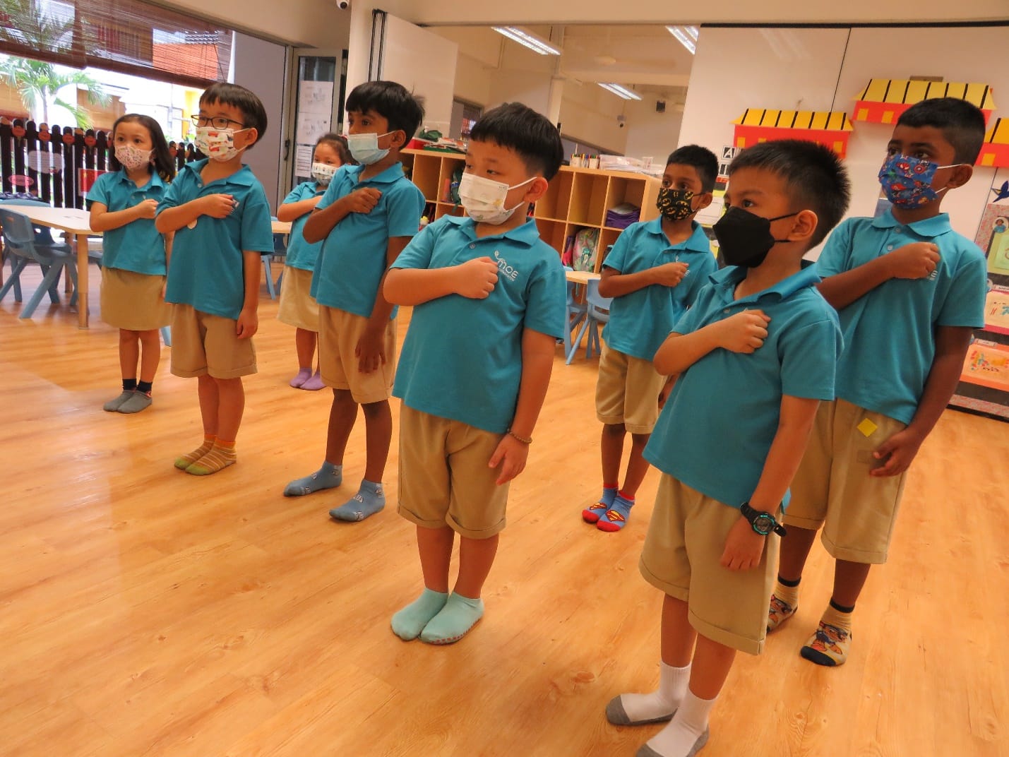 Young children in teal uniforms and face masks stand in rows with hands on chests in a classroom.