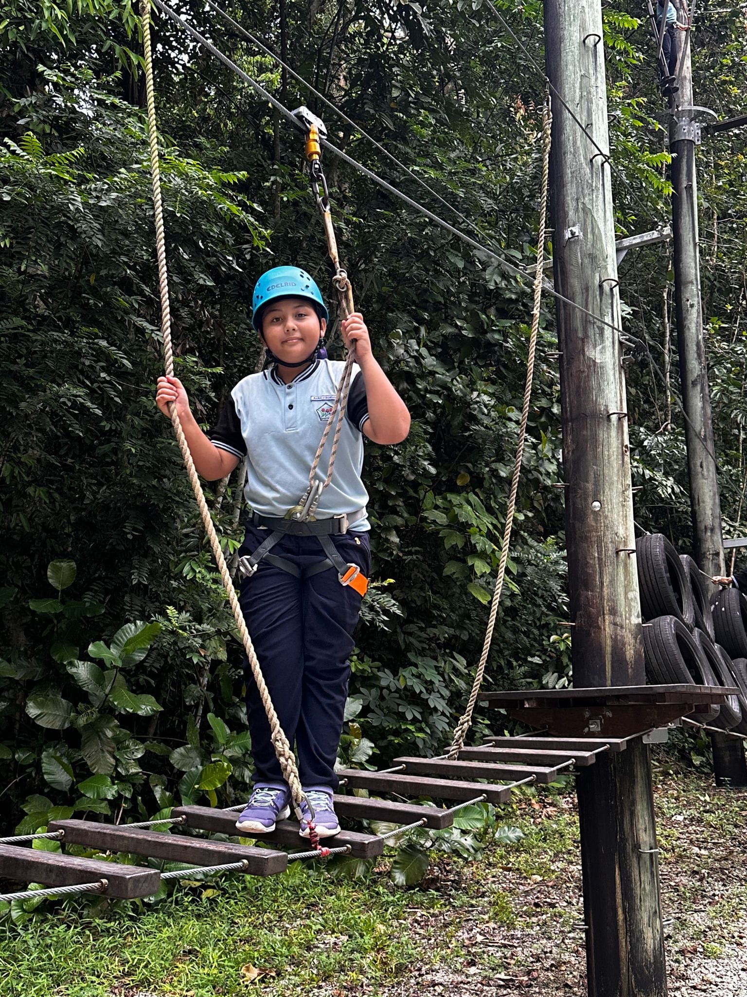 A child wearing a blue helmet and harness crosses a rope ladder obstacle course surrounded by green trees.