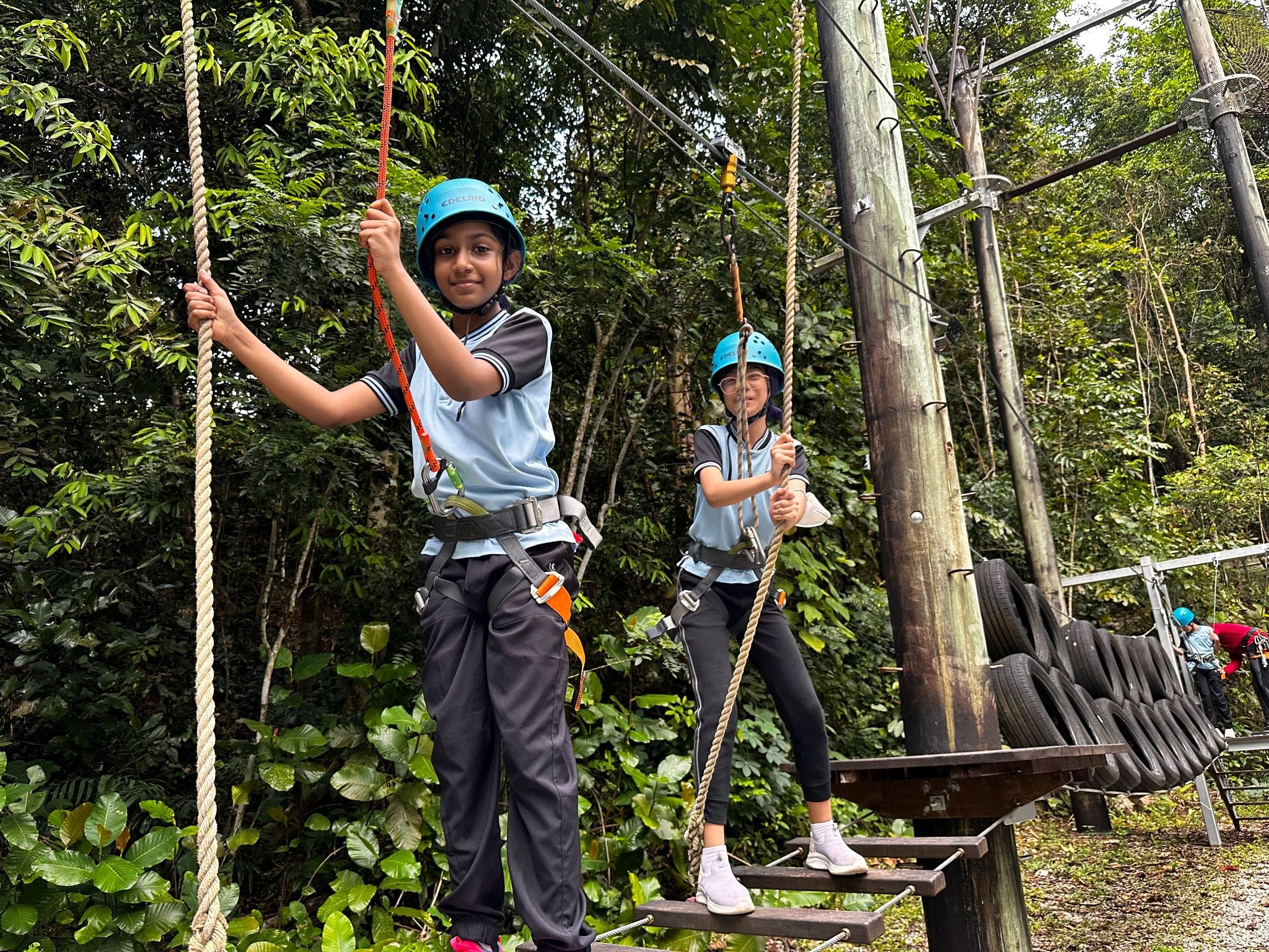 Two children wearing blue helmets and safety harnesses navigate an outdoor rope course surrounded by green trees.