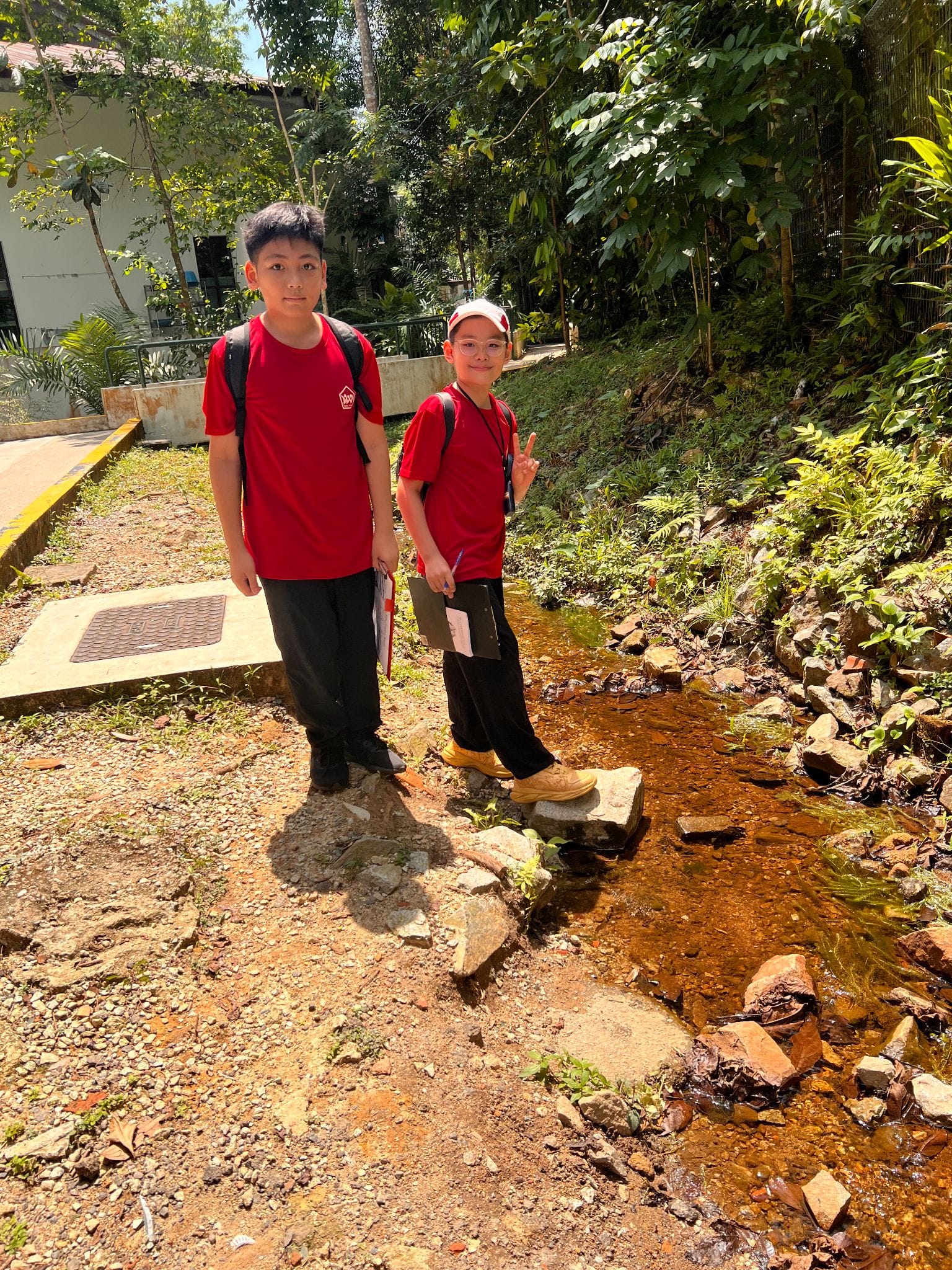 Two boys in red shirts and backpacks stand beside a small rocky stream surrounded by lush green trees.